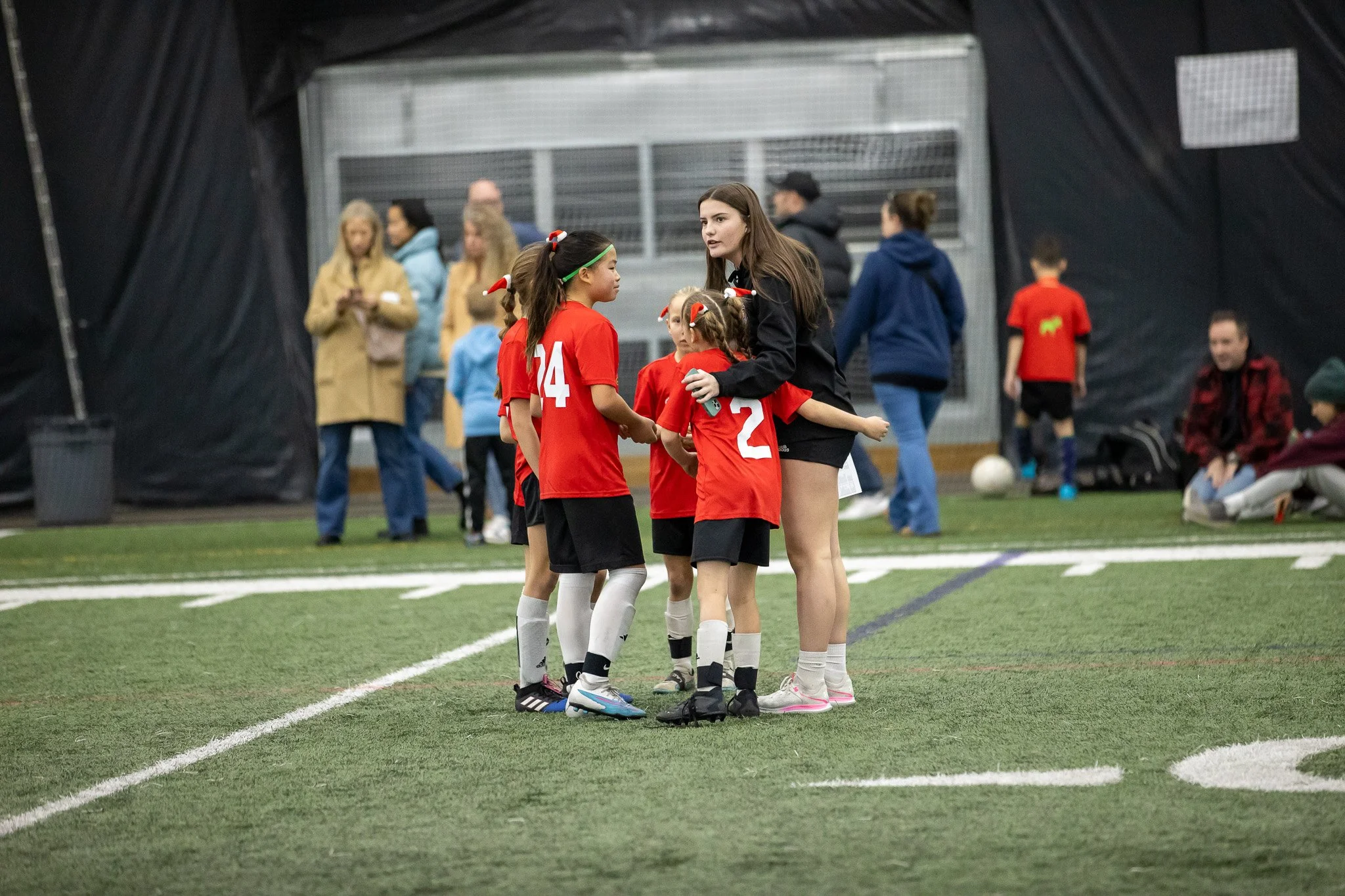 A youth soccer team in red jerseys huddles around their coach during a game inside an indoor field.