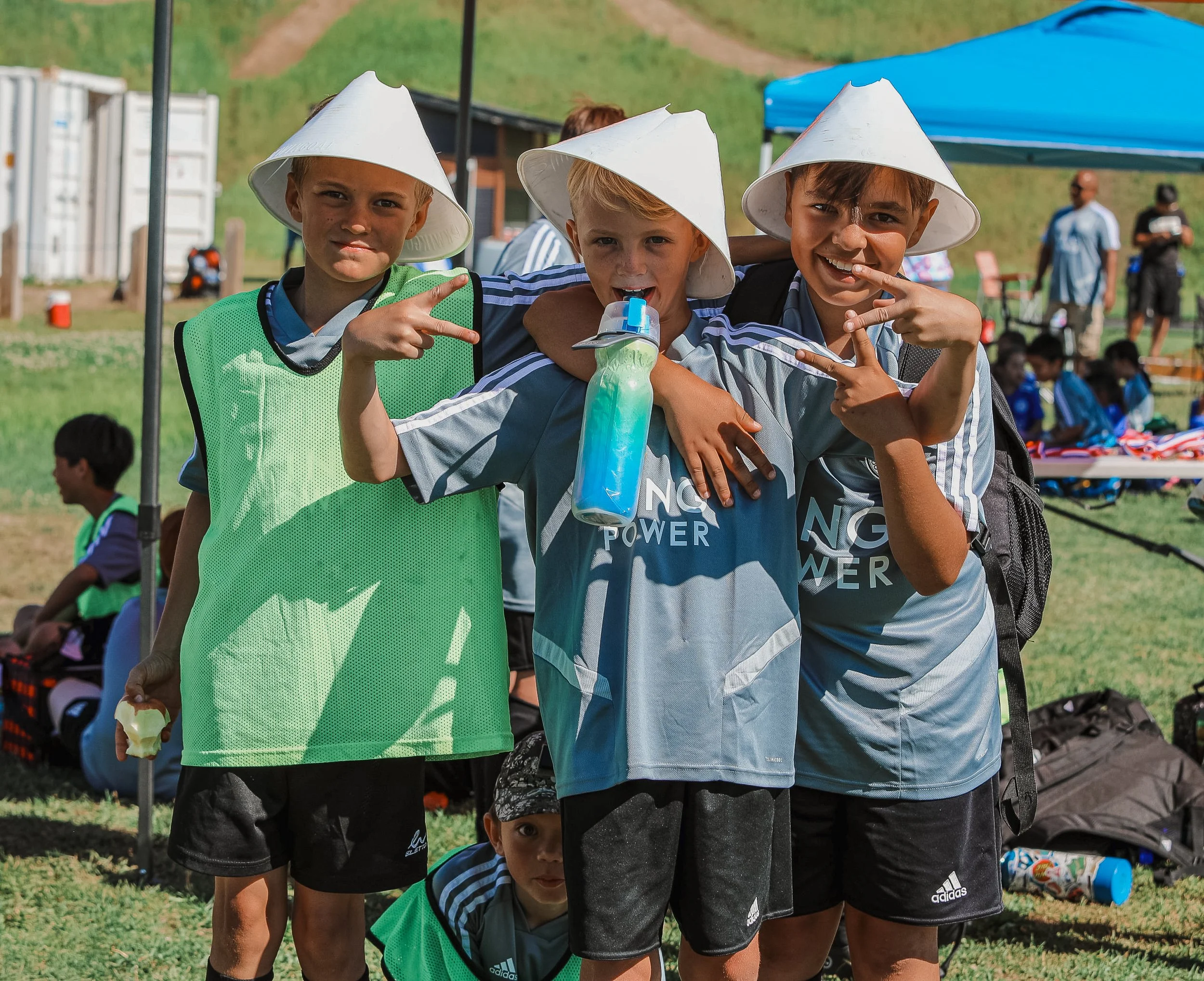 Three young boys in sports uniforms and white hats posing together outdoors during a sports event, with a grassy field and other children in the background.