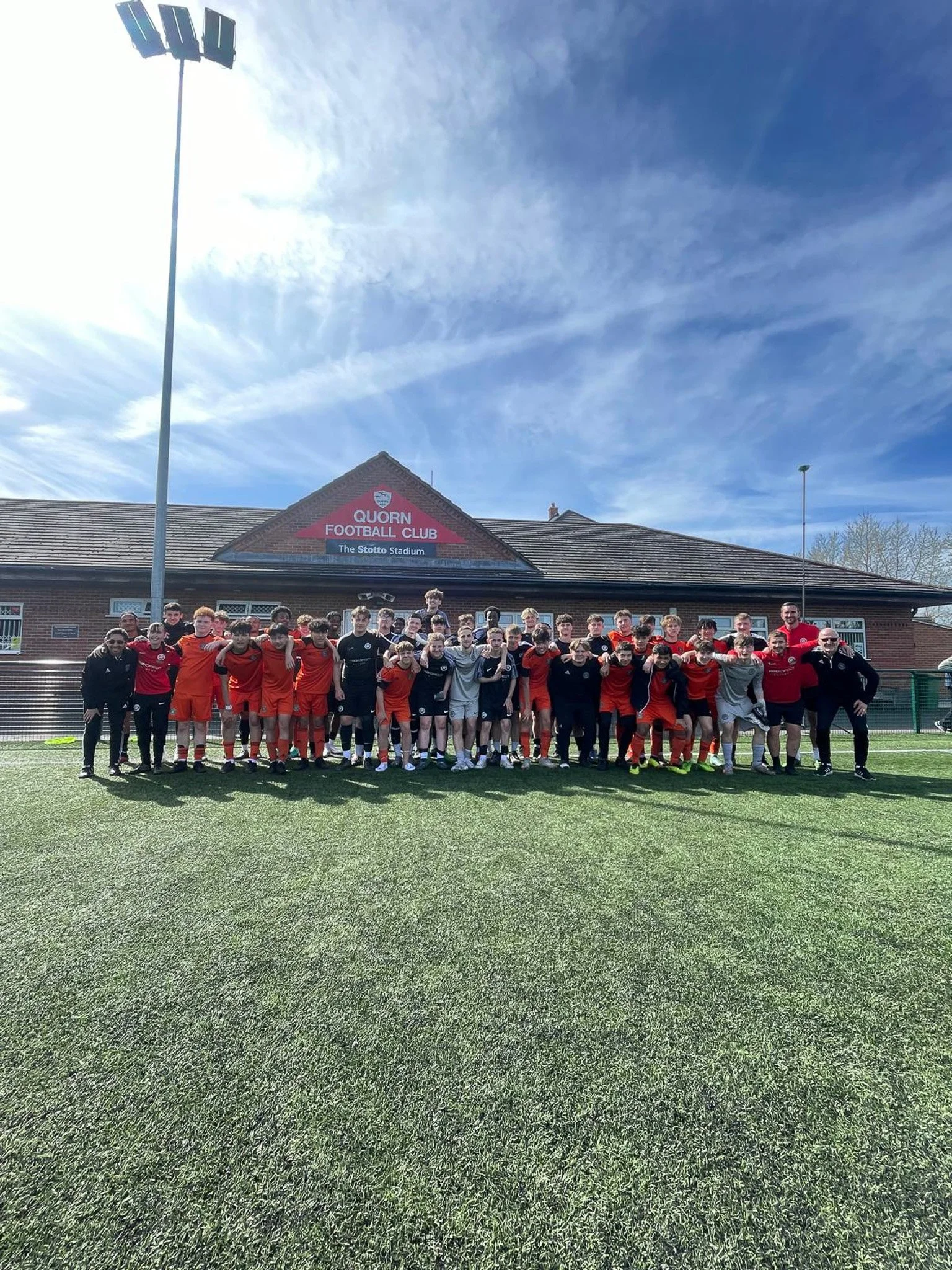 Group photo of a football team and coaching staff on a sports field with a building in the background marked Quorn Football Club and the Stotfo Stadium, under a partly cloudy sky.