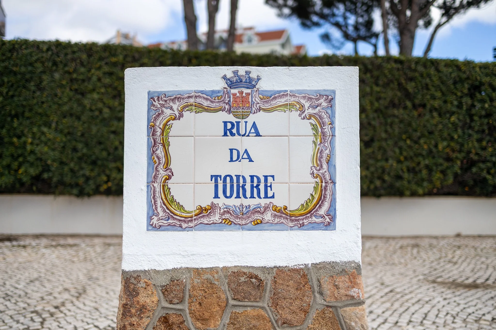 Street sign with decorative border titled 'Rua da Torre' in Portuguese, mounted on a pedestal against a background of a hedge and trees.