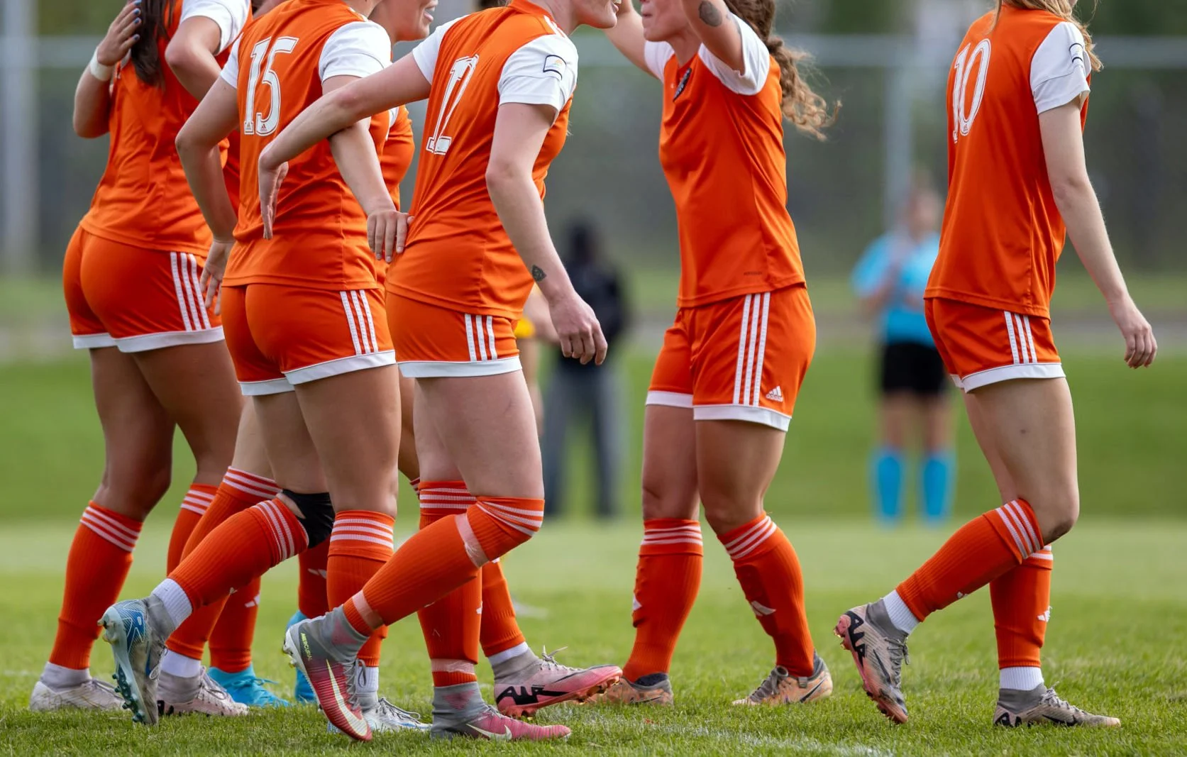 A group of female soccer players in orange and white uniforms standing on a soccer field, appearing to strategize or celebrate during a game.