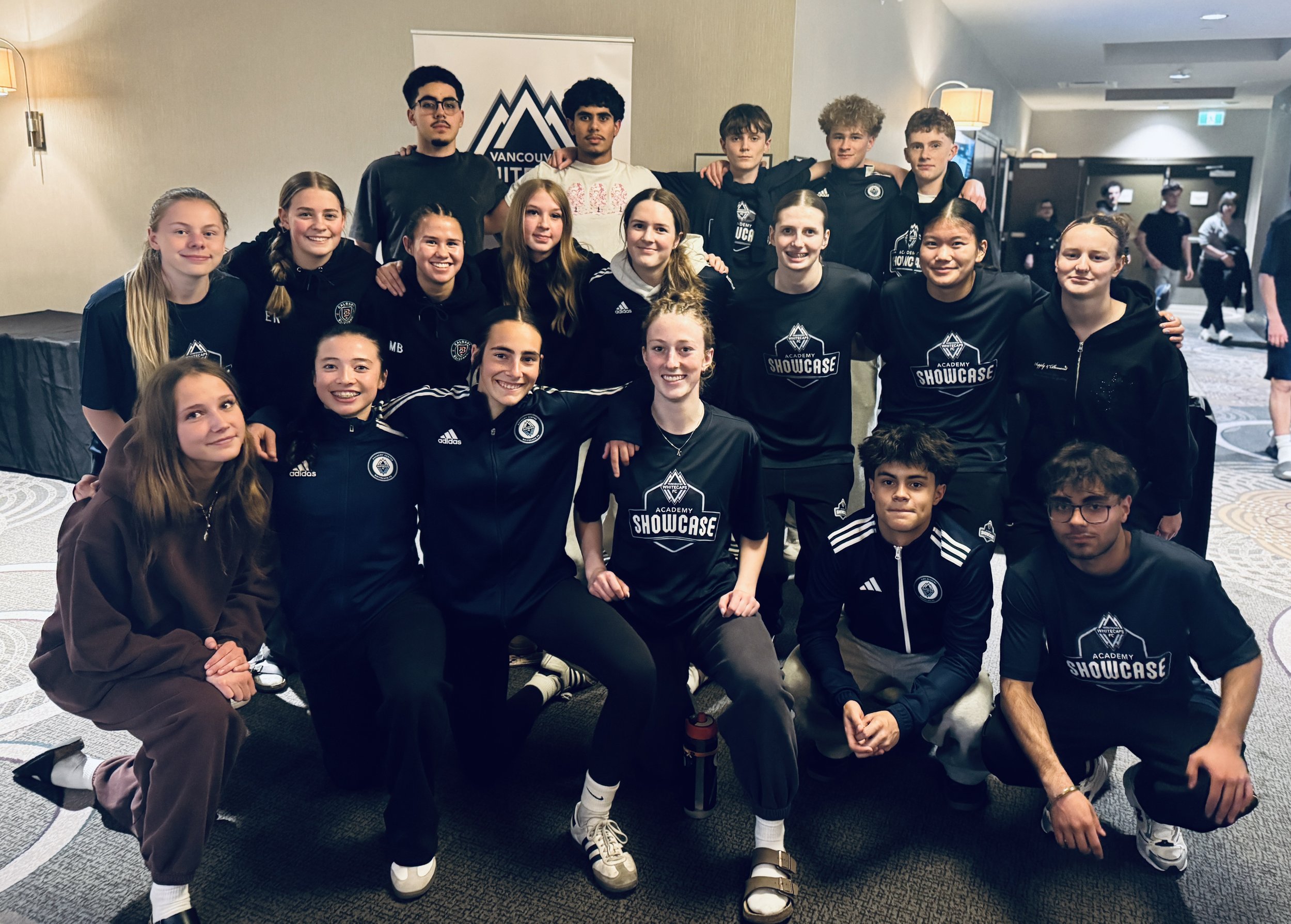 Group of young people posing for a photo indoors, some wearing black athletic clothing with 'Showcase' logos, in front of a Vancouver Whitecaps banner, with others in casual clothes, smiling and standing together.