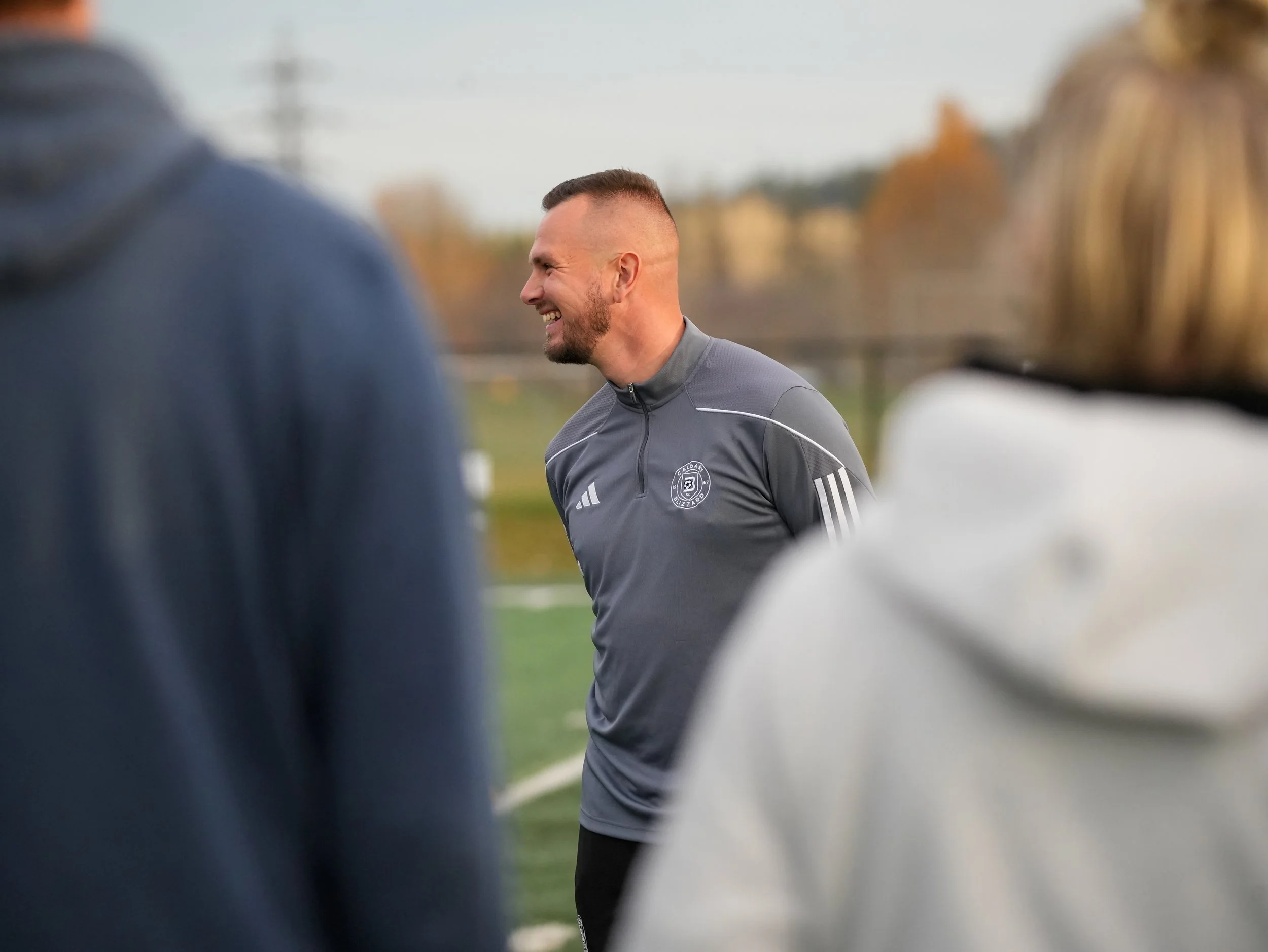 A man in a gray athletic jacket smiling and talking to others on a sports field with trees in the background.