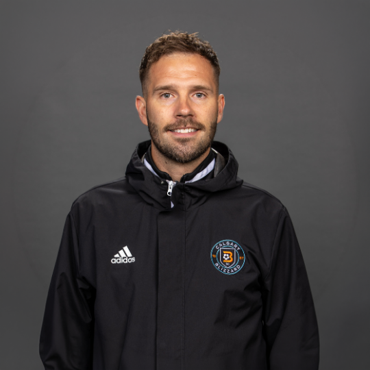 A man with short brown hair and a beard, smiling, wearing a black jacket with a logo on the chest.