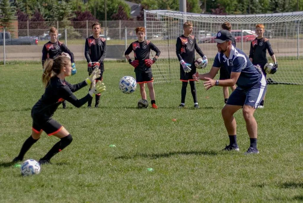 A coach instructs young female soccer players during a practice on a grass field, with a soccer goal in the background.