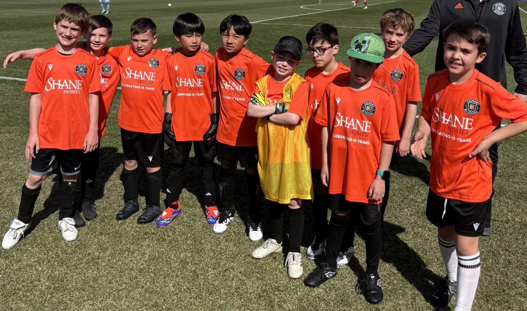 A group of young boys in orange soccer jerseys and black shorts standing on a soccer field. They are posing for a team photo, with some smiling and others looking serious. One boy near the center is wearing a yellow goalkeeper jersey with crossed arm