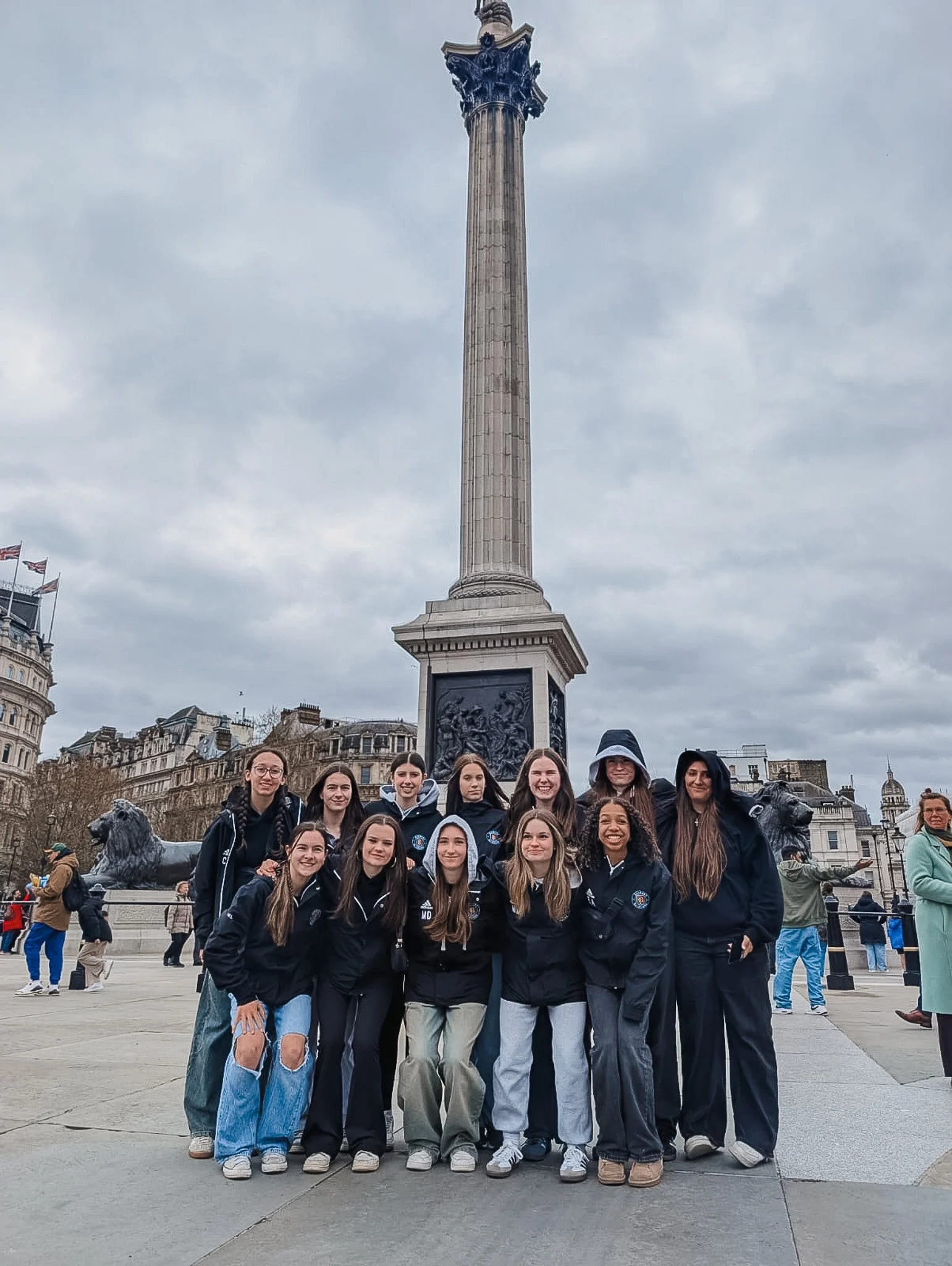 Group of young women posing in front of a tall monument in an urban area with cloudy sky.