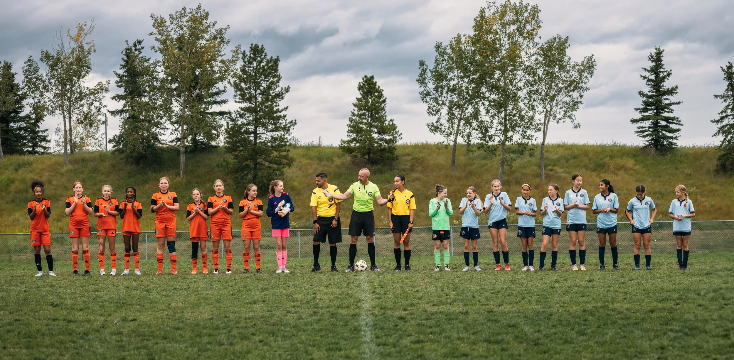 Girls soccer teams in orange and light blue uniforms line up on the field for a pre-game moment, with referees in yellow shirts and a coach in the center.