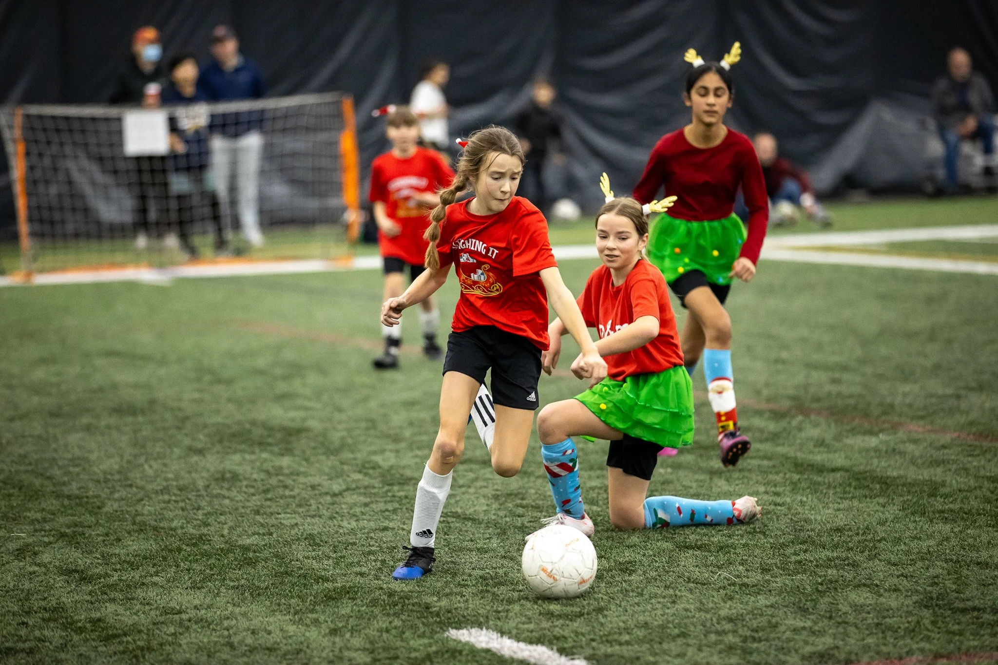 Children playing soccer on an indoor field, dressed in festive holiday costumes with reindeer antlers, some in red shirts and green skirts, during a game.