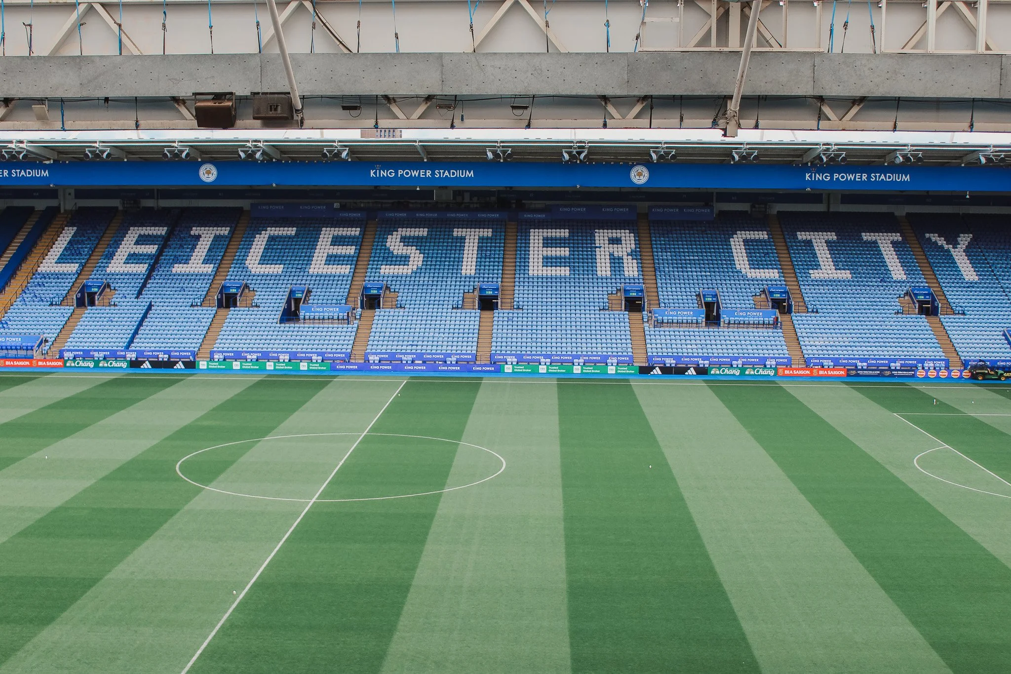 Empty soccer stadium with a field, blue and white seating, and a large sign that reads 'LEICESTER CITY' in the stands.