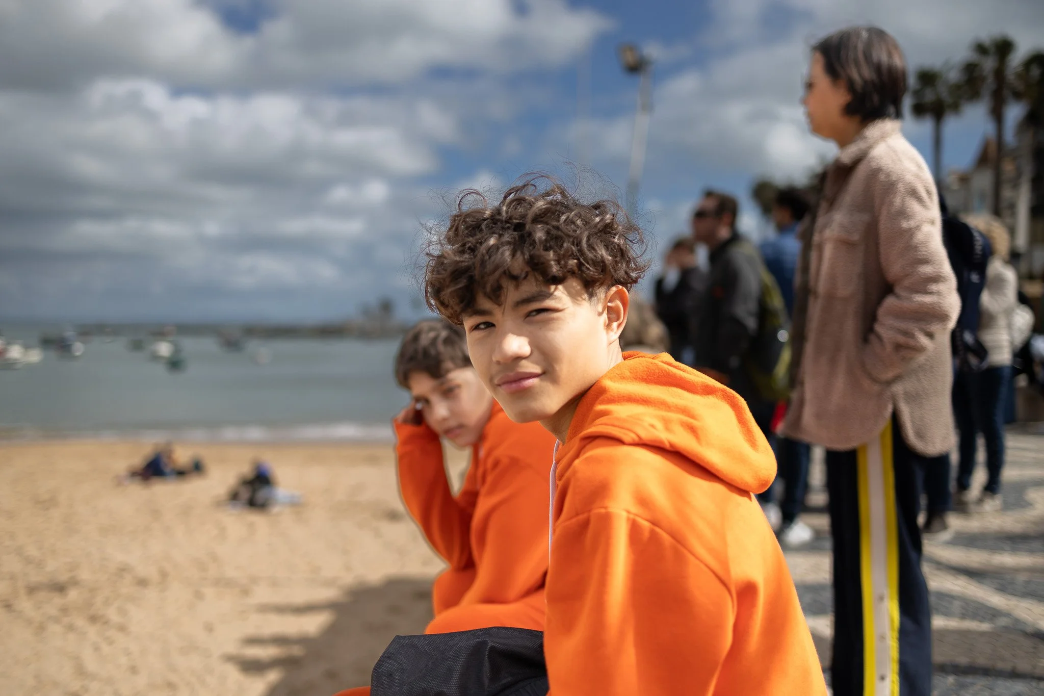 Two boys sitting on a beach, the boy in the foreground has curly hair and wears an orange hoodie, looking at the camera with a slight smile, while the other boy also in an orange hoodie faces away, overlooking the sand and water. In the background, p