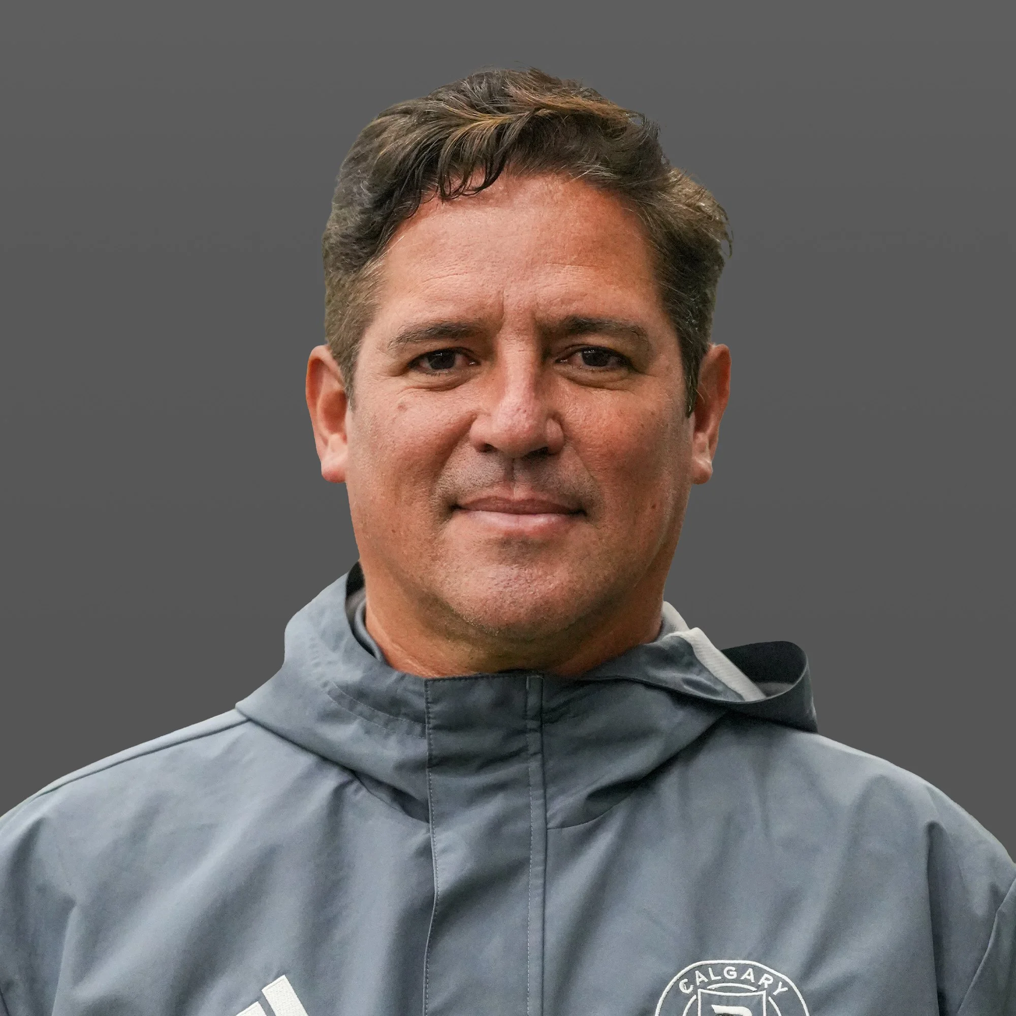 A man in a gray sports jacket stands on a sports field at night, illuminated by bright stadium lights, with a blurred field and players in the background.