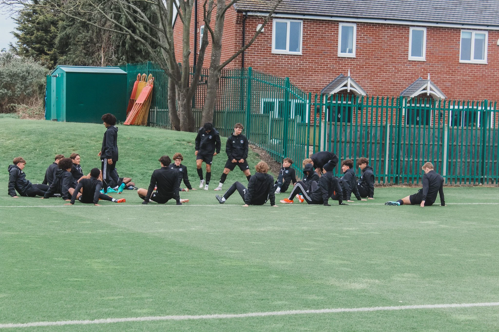 Group of young soccer players sitting and standing on the field during practice or a game, with a fence, trees, and building in the background.