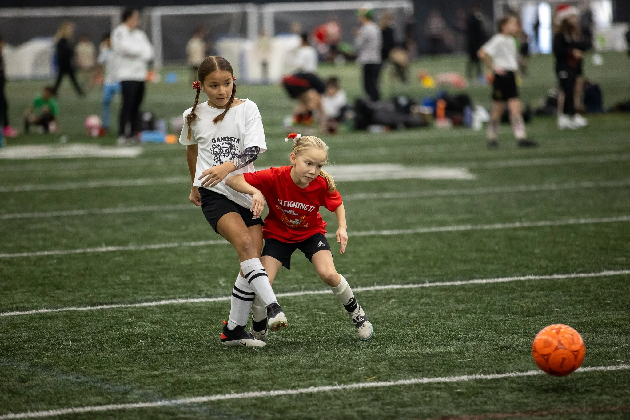 Two young girls playing soccer indoors on a green turf field. One girl in white and black sportswear is chasing an orange soccer ball as the other girl in red attempts to kick it. Other children and adults are visible in the background.