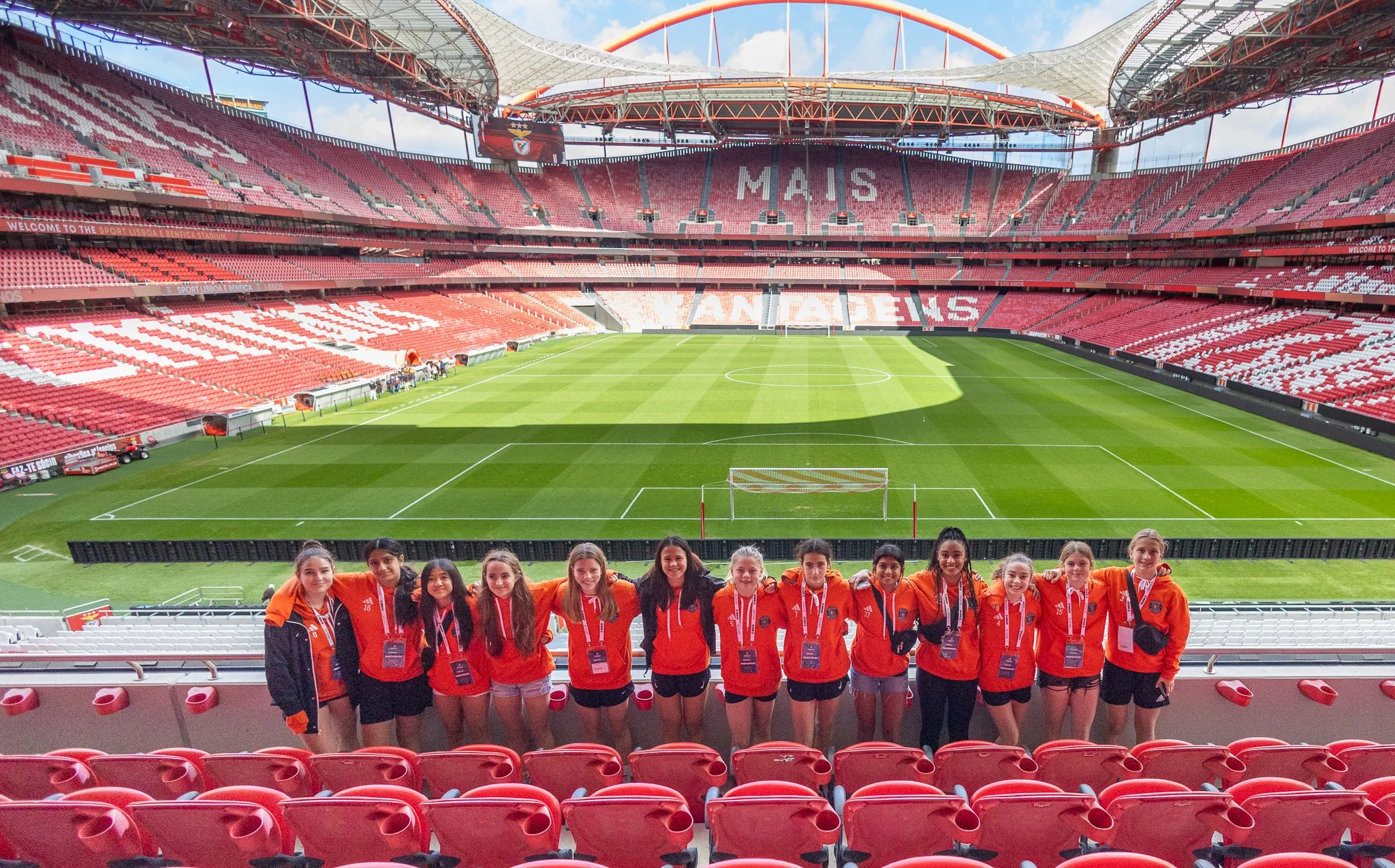 A group of young women wearing orange team jackets and medals standing in front of a soccer field inside a large stadium with red seats and a partially covered roof.