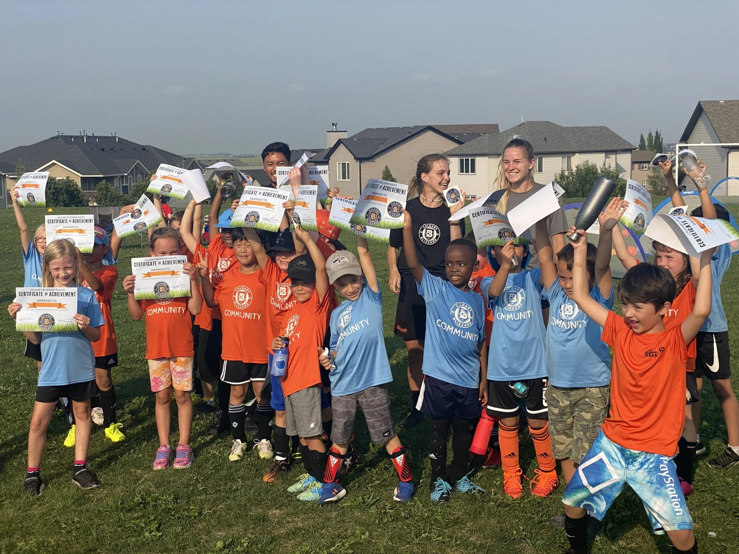 Group of children and adults celebrating soccer victory with certificates and trophies on a grassy field, with residential houses in the background.