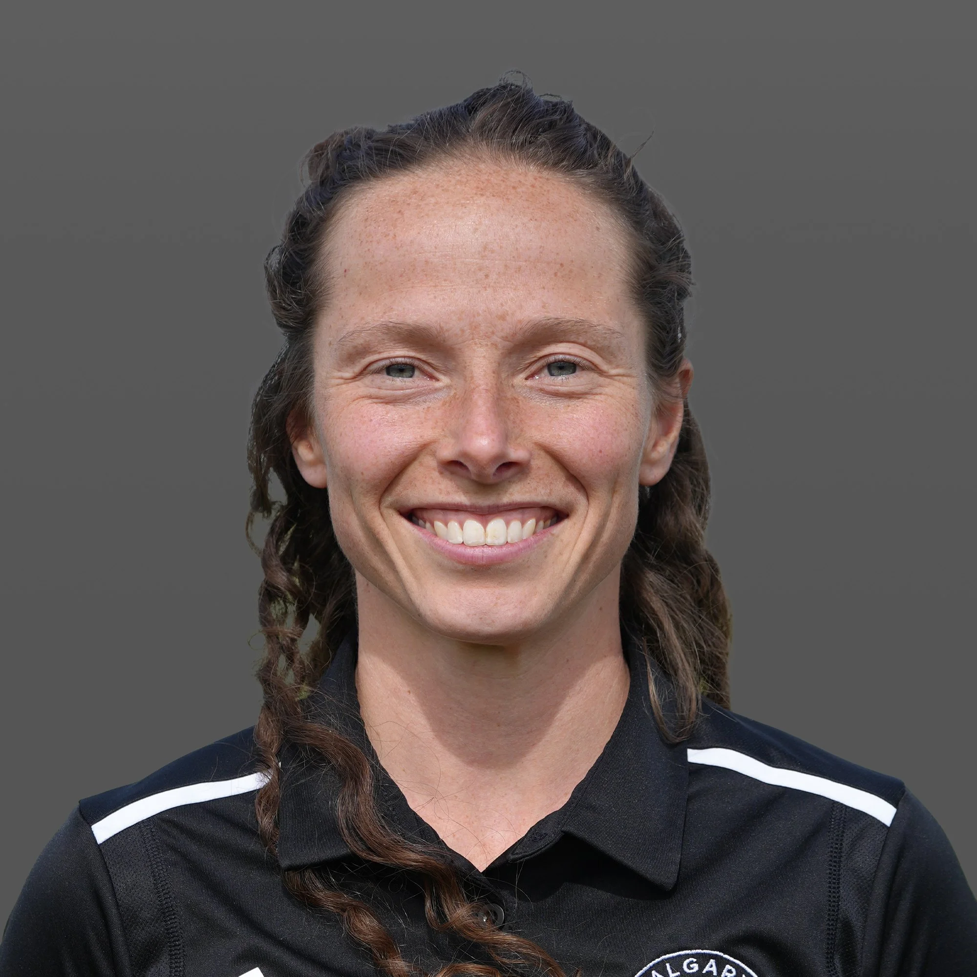 Close-up of a smiling woman with blue eyes, freckles, and curly brown hair pulled back, wearing a black athletic jacket with white stripes, standing outdoors on a grassy field during daytime.