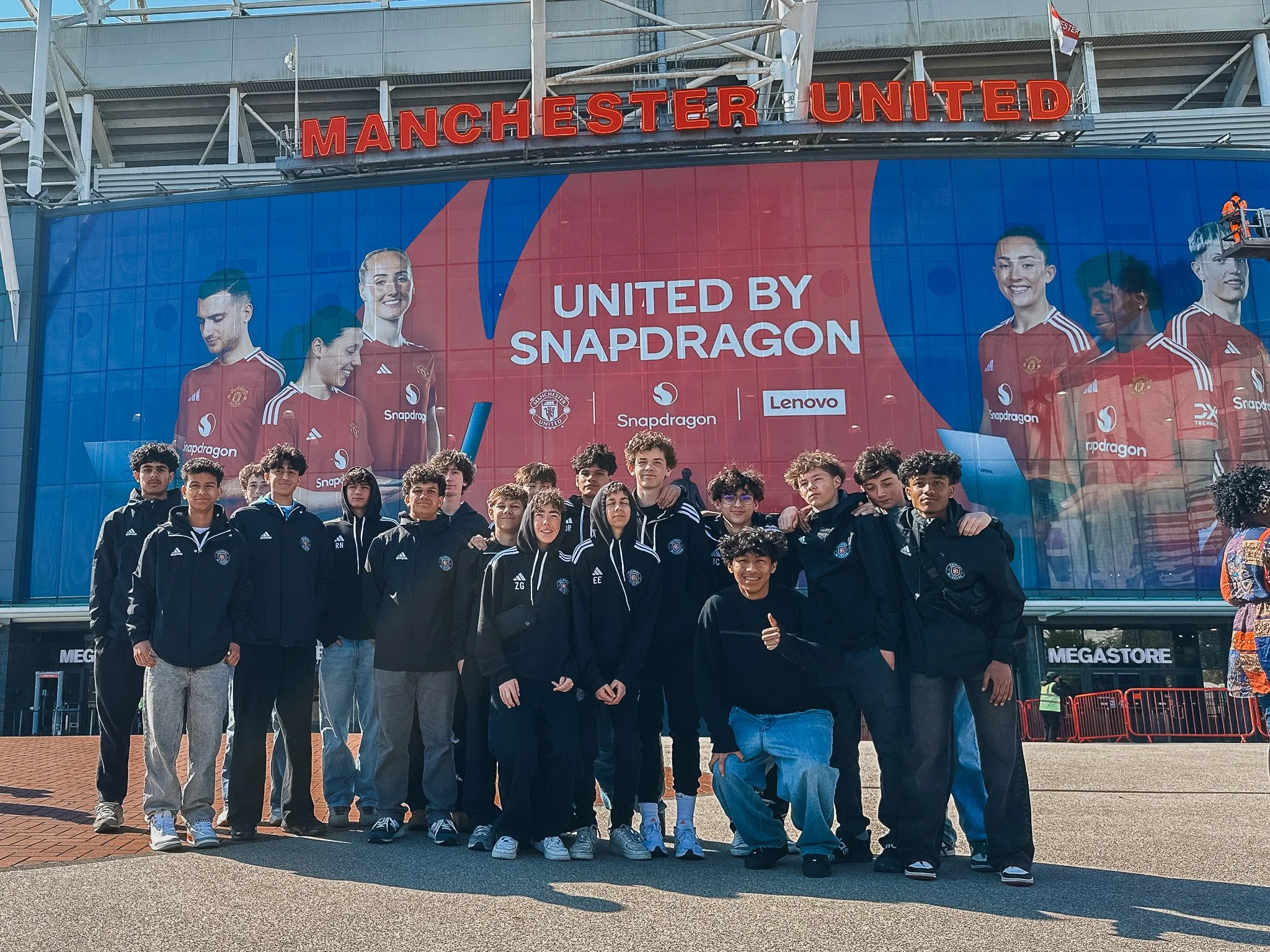 A group of young men standing in front of Manchester United stadium's exterior. Large banner behind them reads 'United by Snapdragon,' featuring images of female athletes in red jerseys. The stadium signage displays 'Manchester United' in red lights.