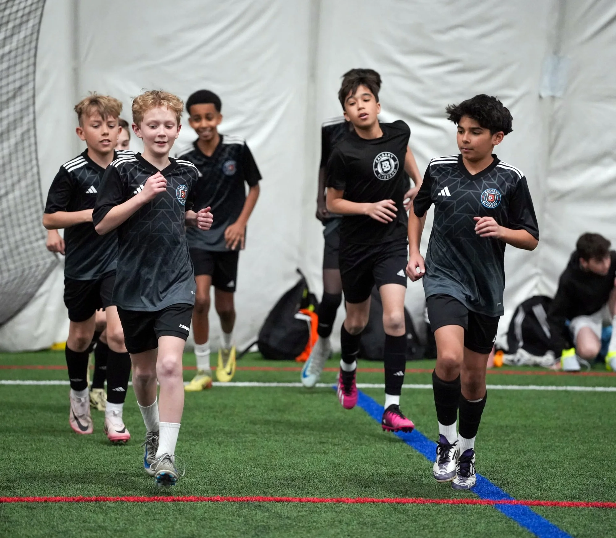 Group of young boys running indoors on a synthetic turf field, wearing black soccer uniforms with a logo, during a soccer practice or game.