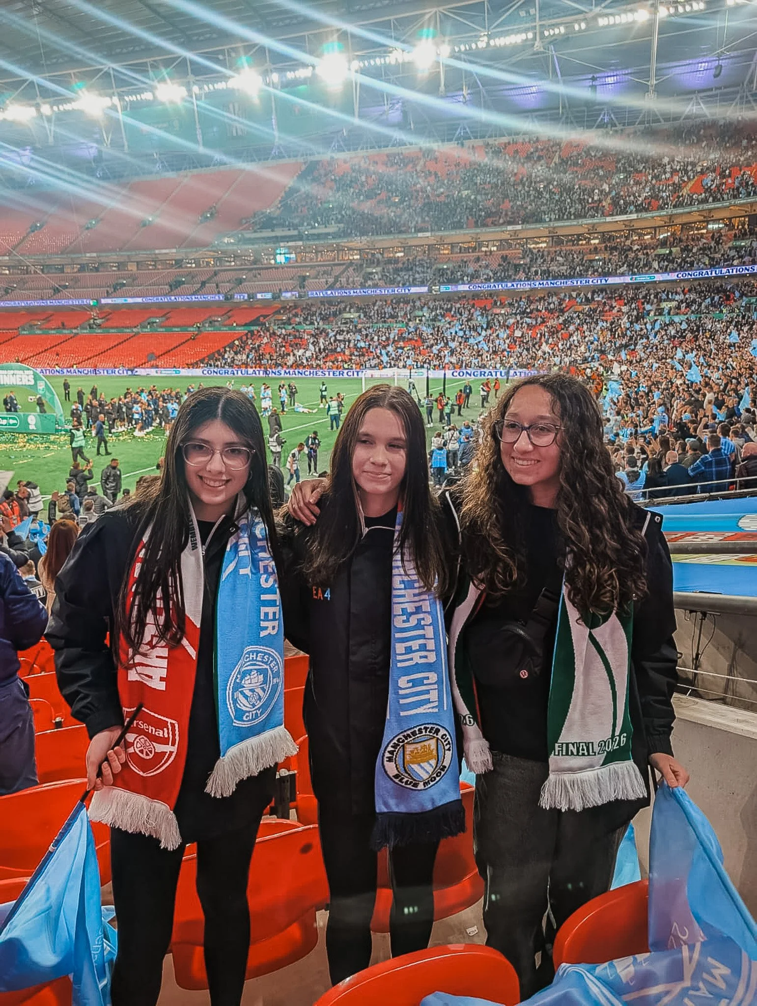 Three young women standing in a stadium, smiling, dressed in jackets and scarves supporting Manchester City and Arsenal, watching a football match on the field.