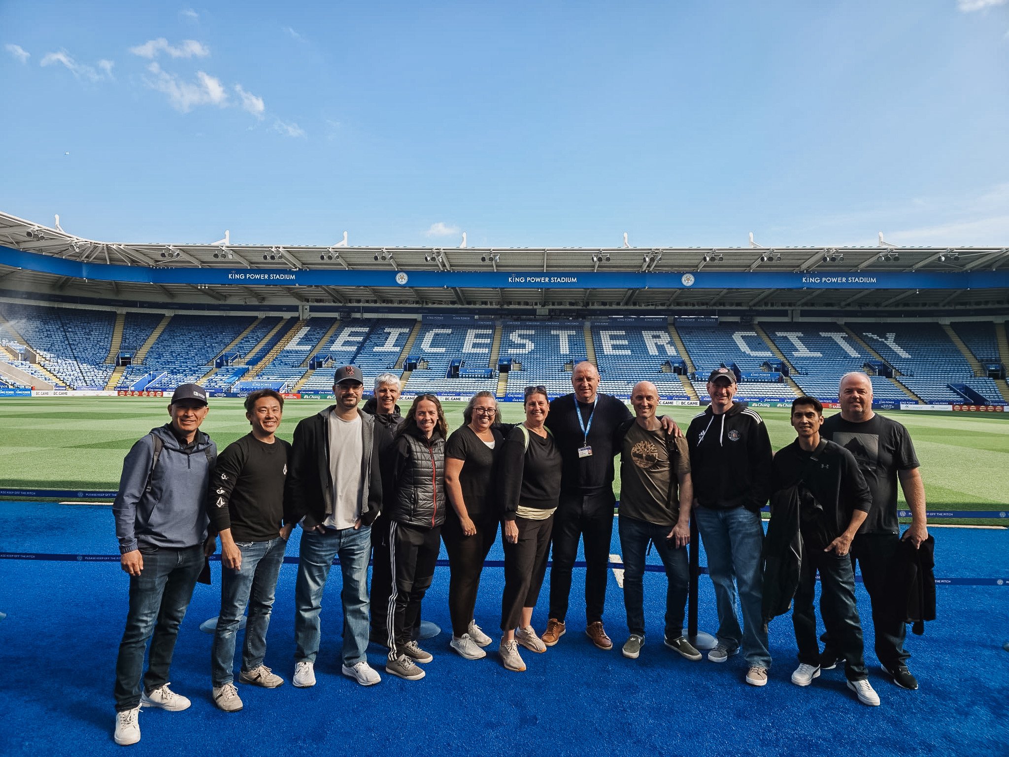 Group of people standing on a blue track at King Power Stadium, Leicester City football stadium, with the stadium seats and field in the background.