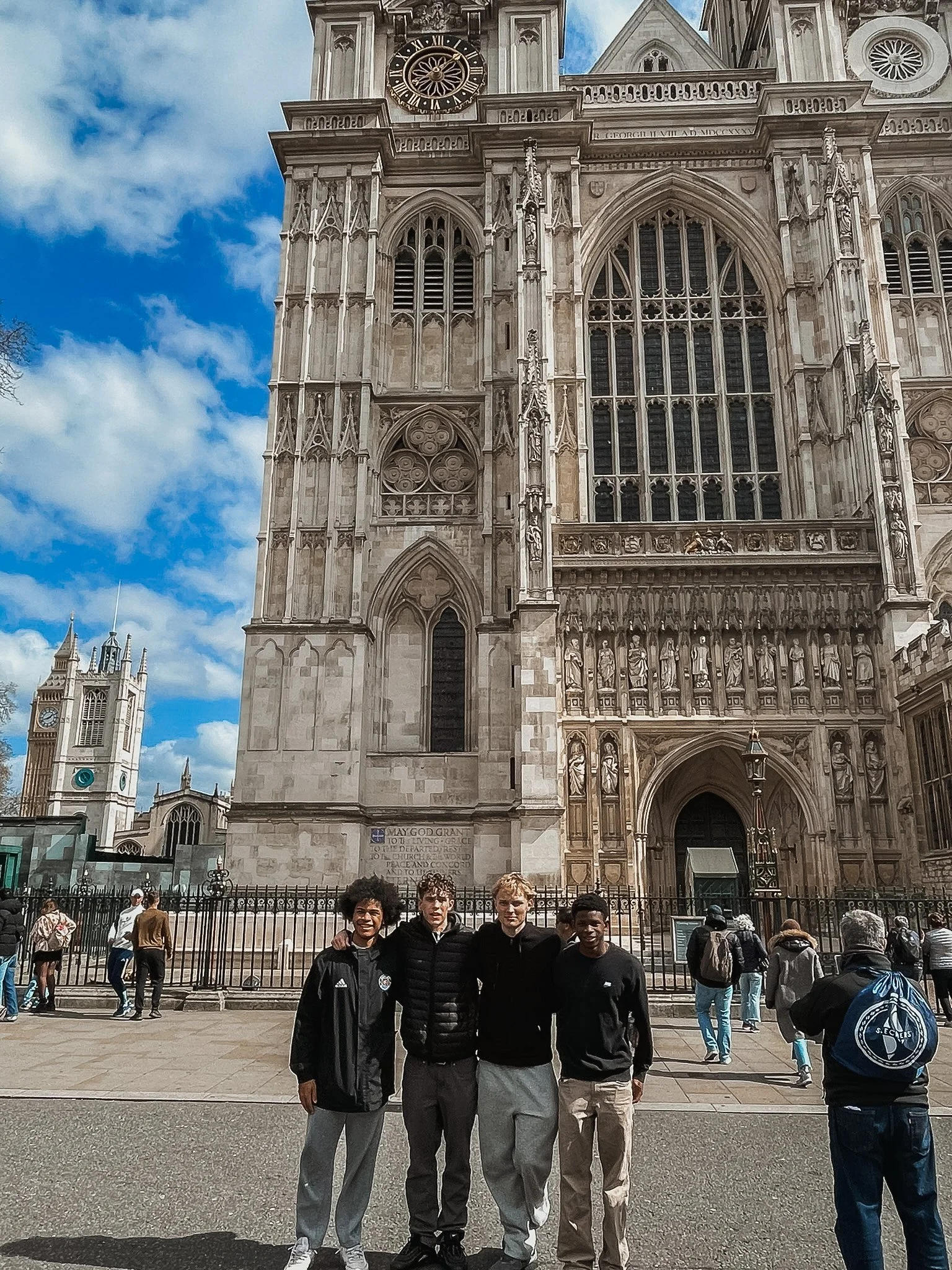 Four young men standing in front of Westminster Abbey in London, with the abbey's Gothic architecture and clock tower visible in the background. Several other people are walking around in the area.