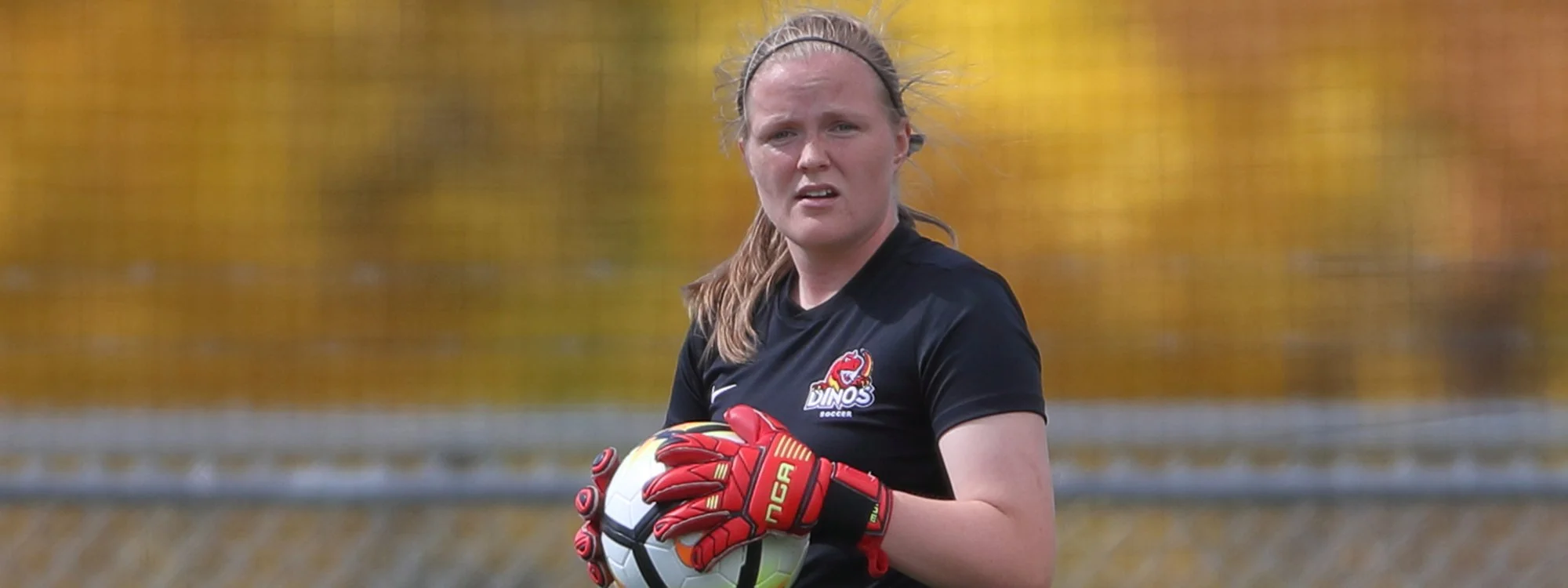 A female soccer goalkeeper holding a soccer ball, wearing red gloves, black jersey with a Dino's Soccer logo, and standing on a soccer field with a yellow and green background.