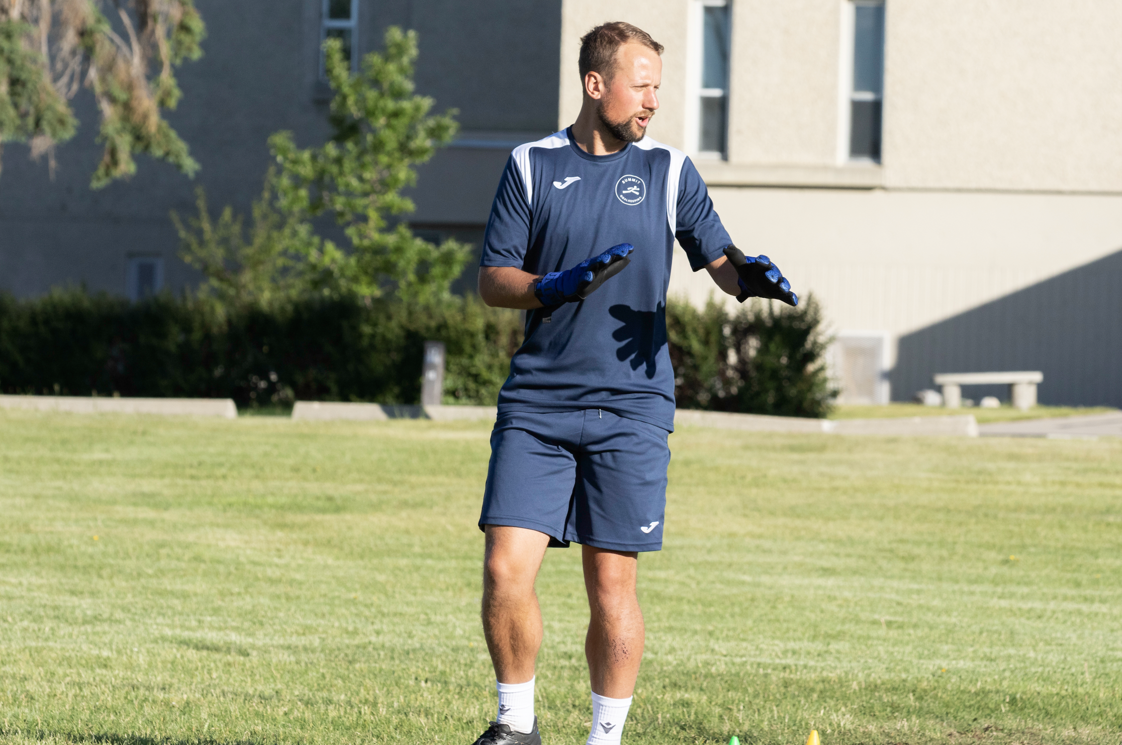 A man in sports gear, wearing a navy blue jersey and shorts with gloves, standing on a grassy field during daytime.