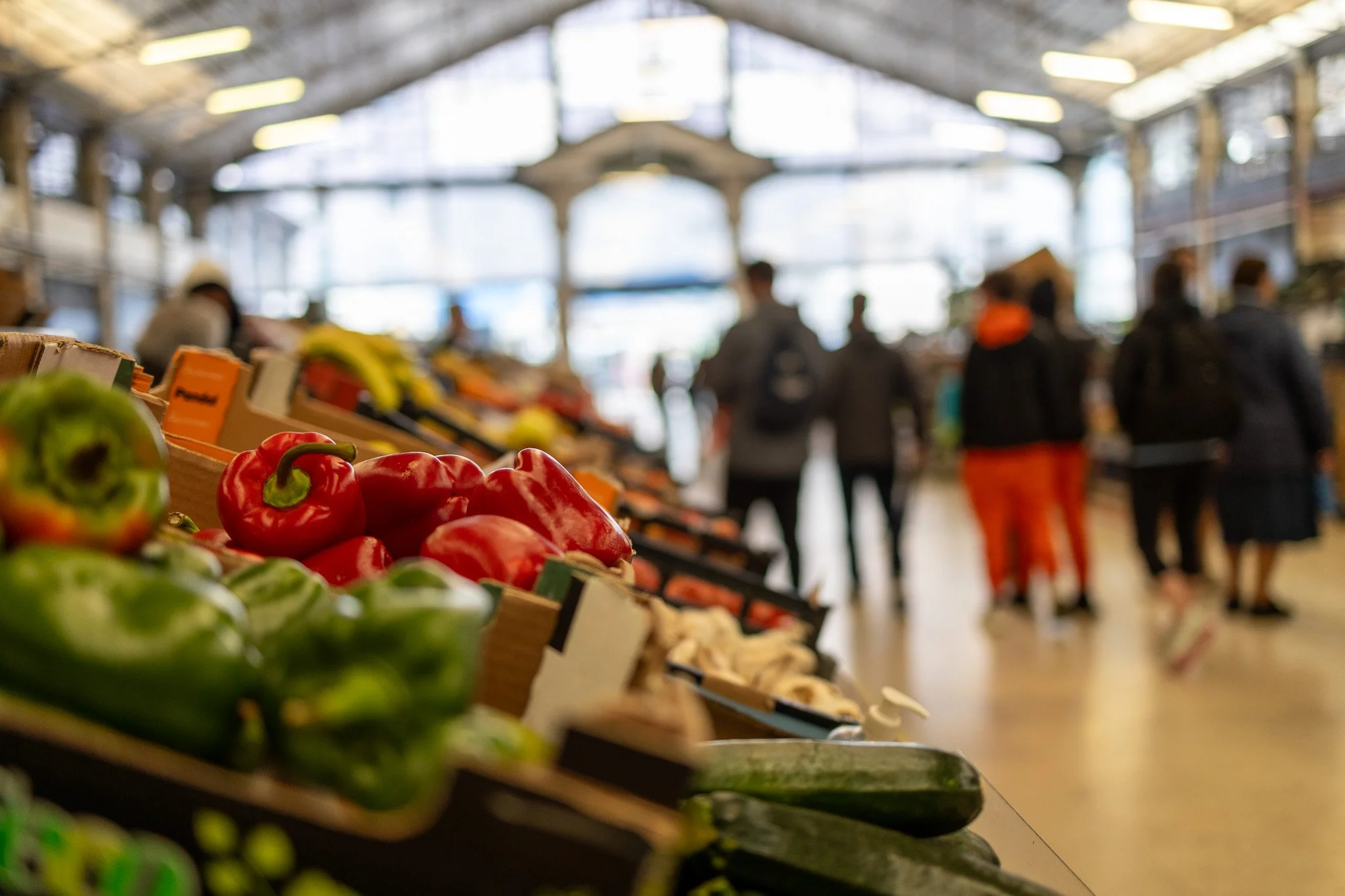 Produce section with red and green peppers at a market or grocery store, with shoppers in the background inside a large building with high glass ceilings.