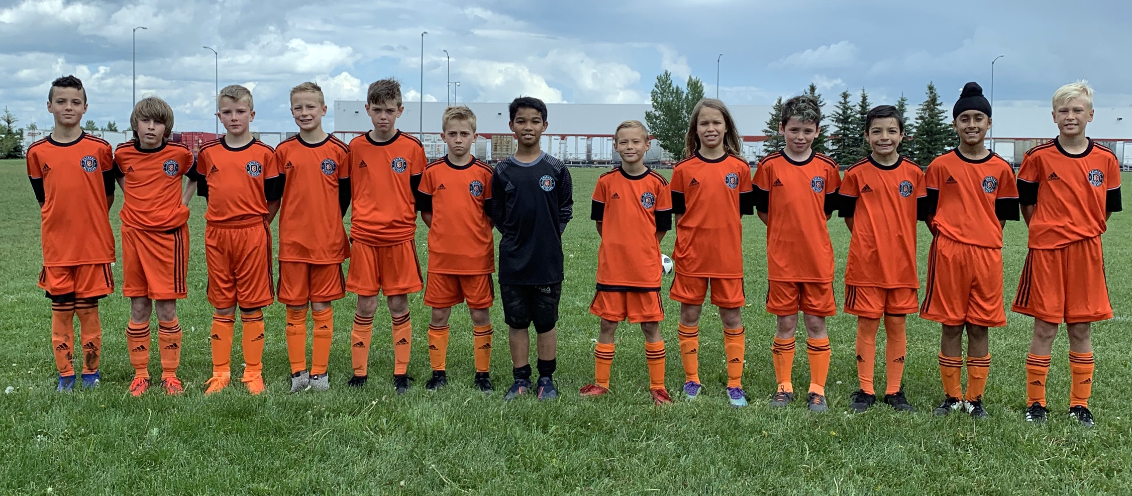 Children in orange soccer uniforms standing in a row on a field with green grass, trees, and a cloudy sky in the background.