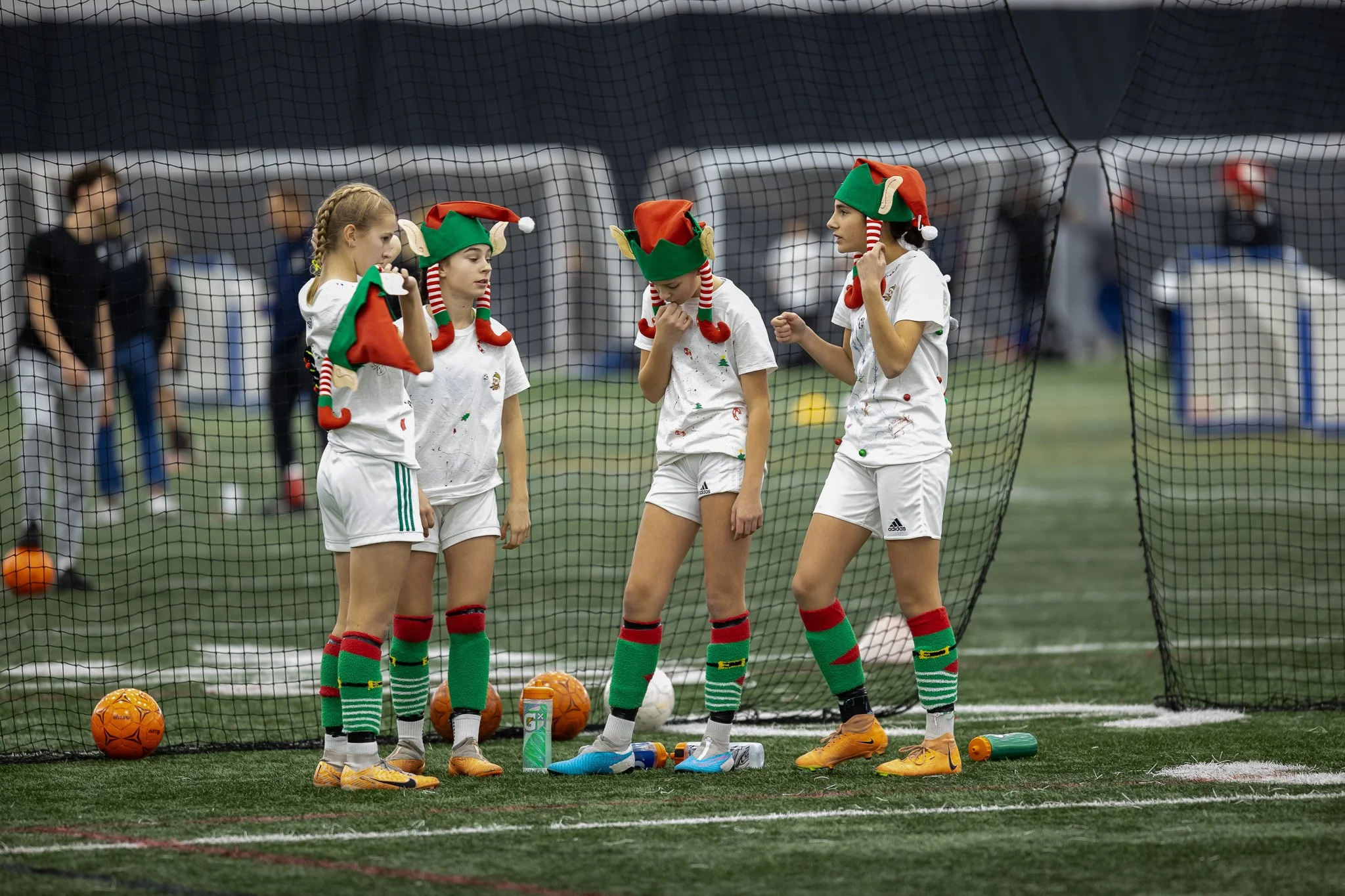 Four young girls in white sports uniforms and Christmas elf hats stand behind a soccer goal net, preparing for a game on an indoor field.