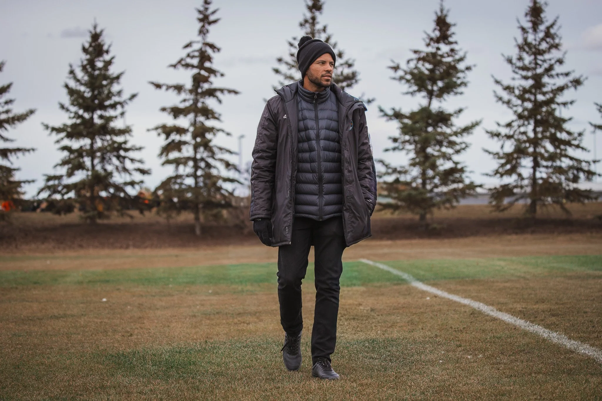 Man walking on a grassy field with tall trees in the background, dressed in a black winter jacket, black pants, gloves, and a beanie.