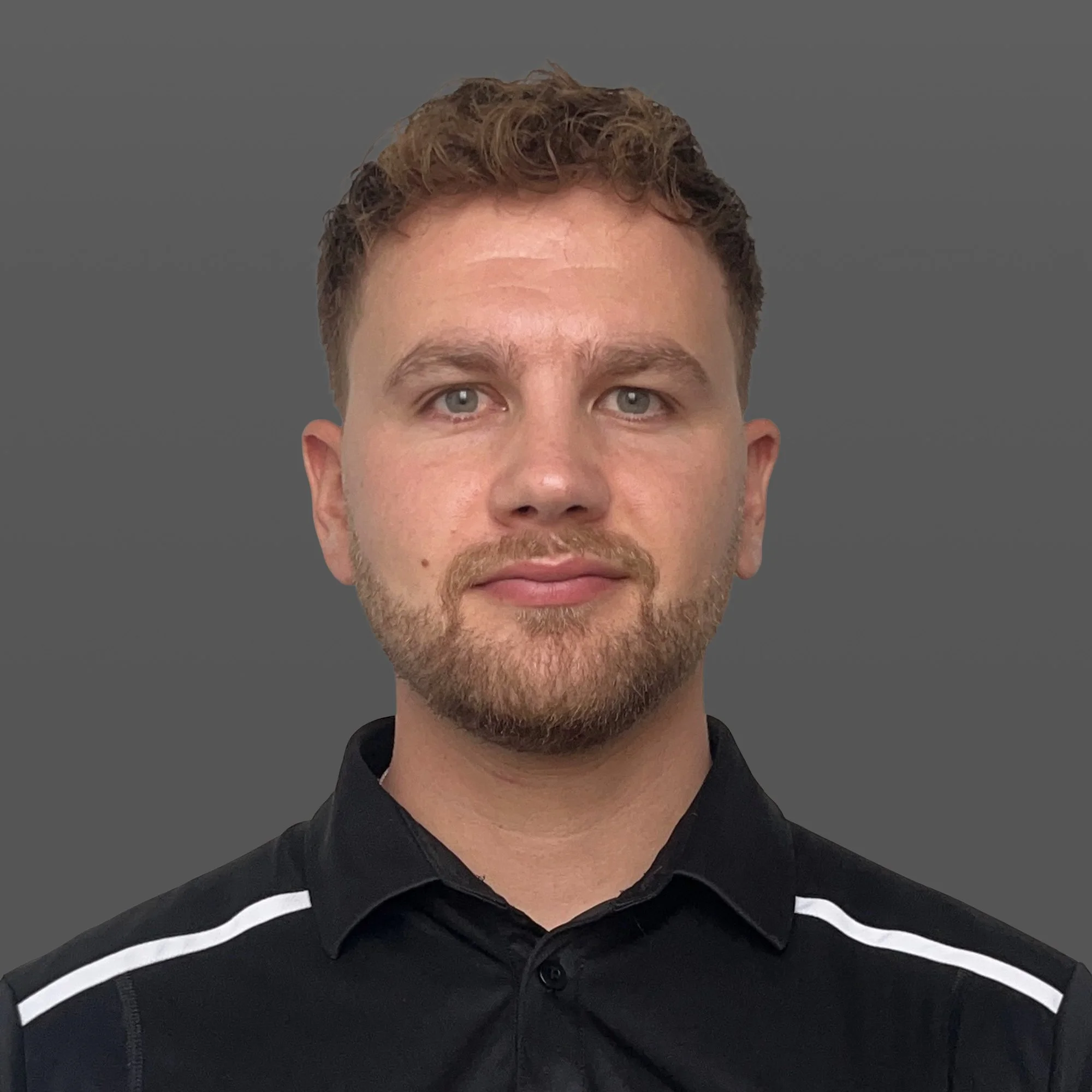 A young man with curly brown hair and a beard stands on a soccer field at night, wearing a black sports jacket with the Adidas logo and a soccer team crest.