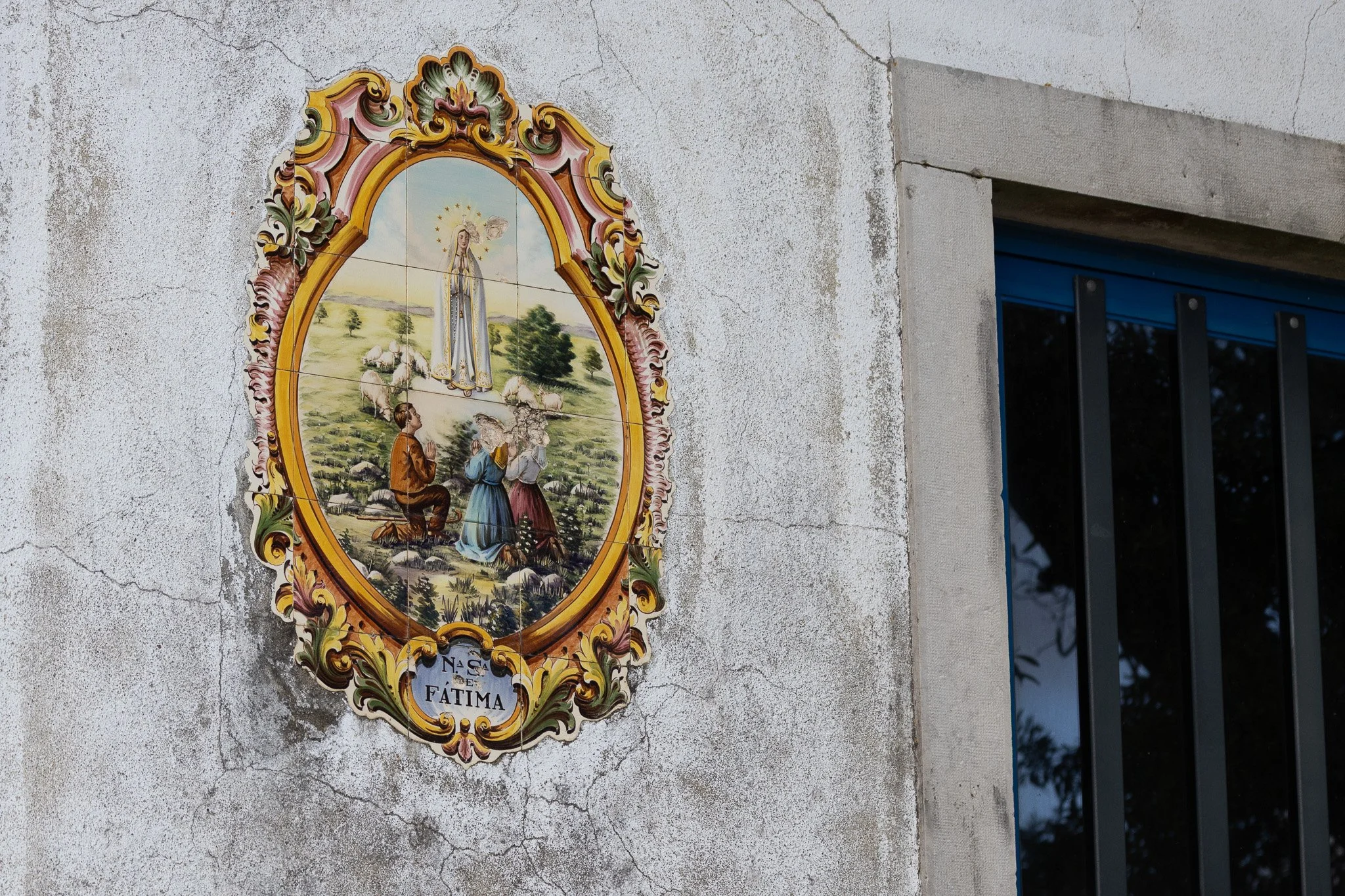 Religious tile mural depicting children praying before a statue of the Virgin Mary, with the sign 'N.S. Fatima' below the image, mounted on a weathered concrete wall near a window with blue frame and black bars.