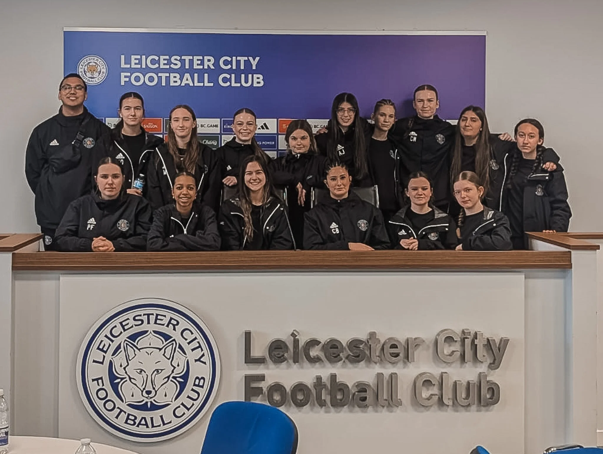 Group of women and men in black Leicester City Football Club jackets posing for a team photo behind a desk with the club's logo and name.