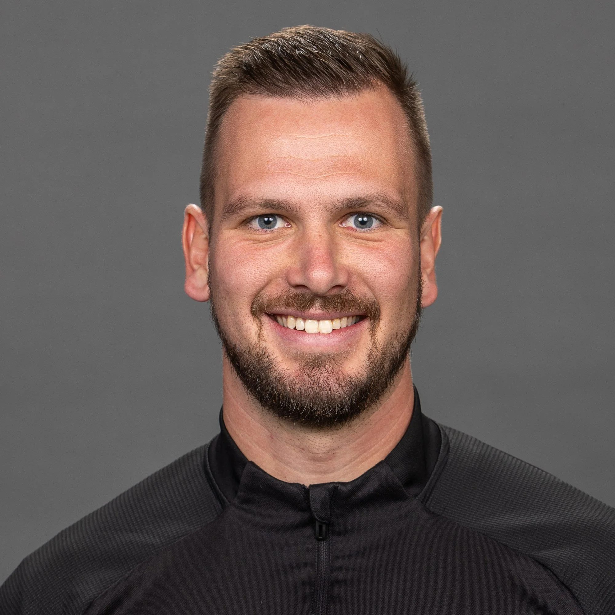 A young man with short brown hair and a beard, wearing a black Adidas sports jacket with a logo that reads "HIGGS." He is standing outdoors at night with a blurred background of a sports field.