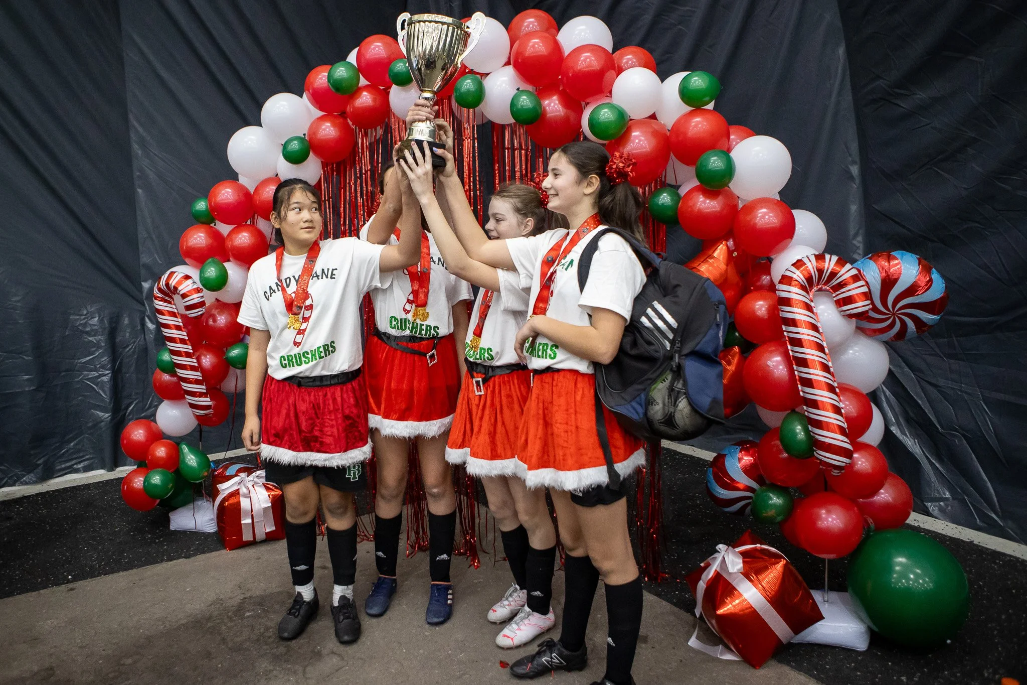 Four young girls in sports uniforms celebrating and holding up a trophy, surrounded by Christmas-themed decorations including balloons, candy cane props, and gift boxes.