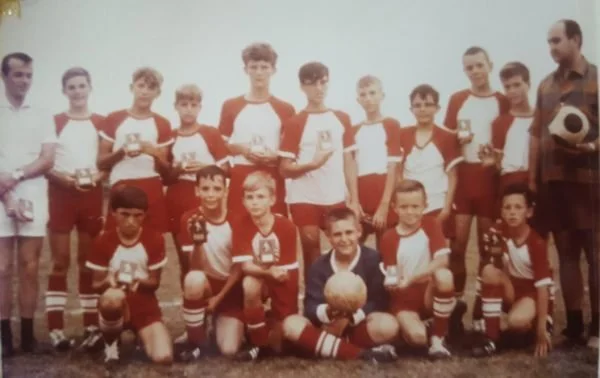 Group of young soccer players and coach posing on a field, holding trophies and a ball, wearing red and white uniforms.