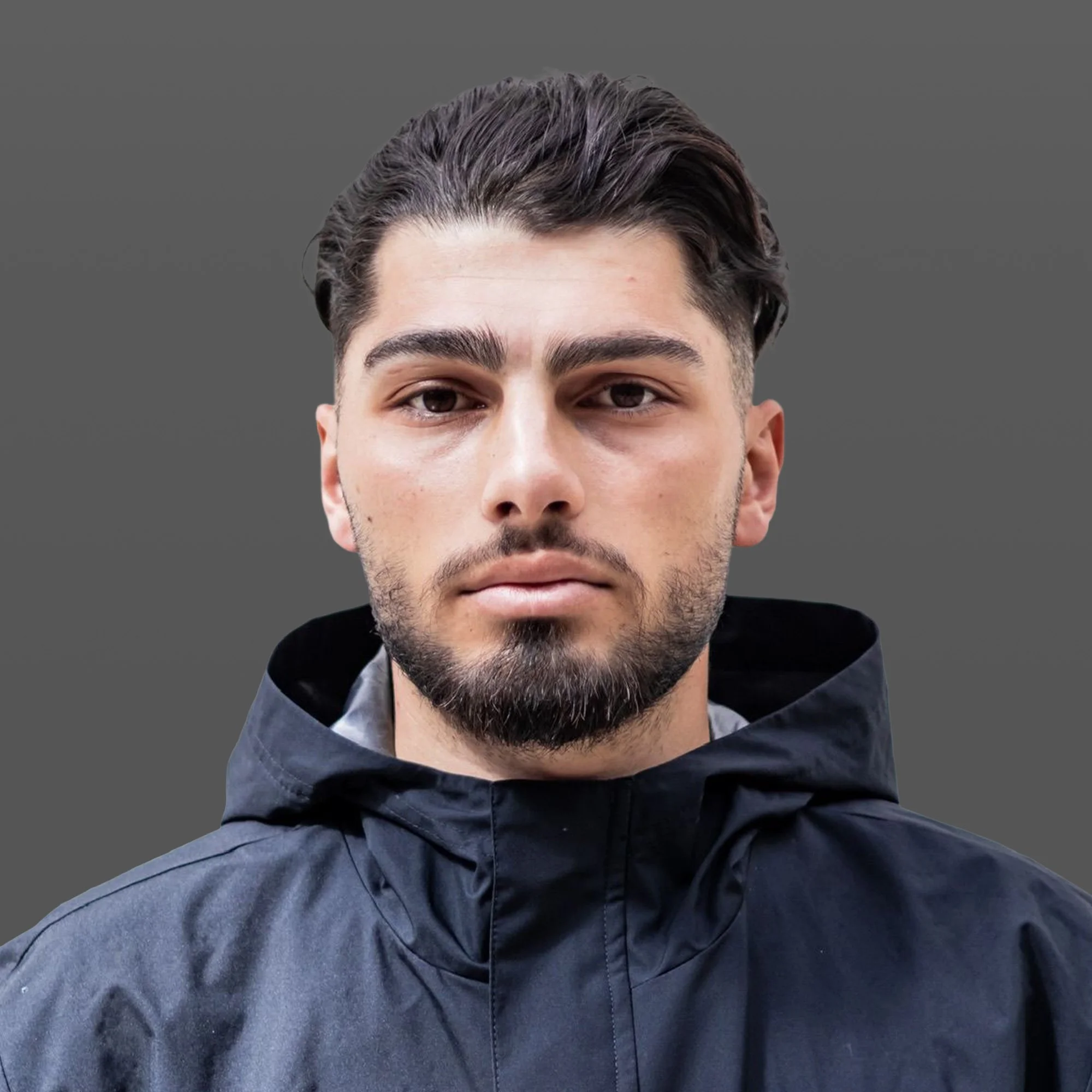 A young man with dark hair and a beard standing outdoors at night, wearing a black hooded jacket, looking directly into the camera with a serious expression.