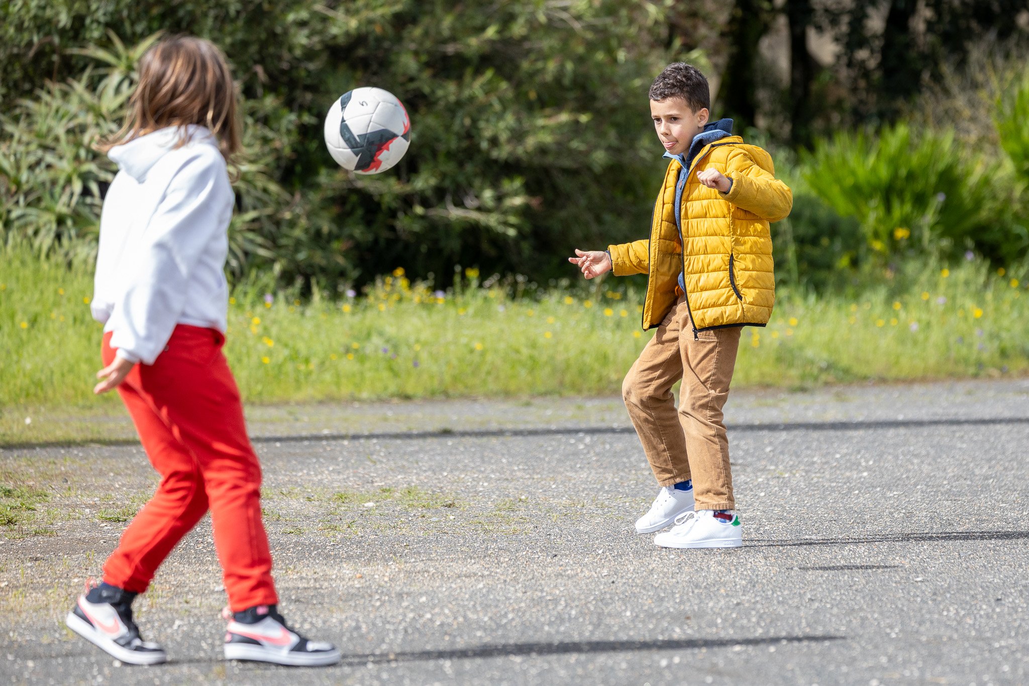 Two kids playing soccer outdoors on a paved area. One kid, wearing a white hoodie and red pants, faces away. The other kid, wearing a yellow jacket, brown pants, and white sneakers, is positioned to kick the soccer ball.