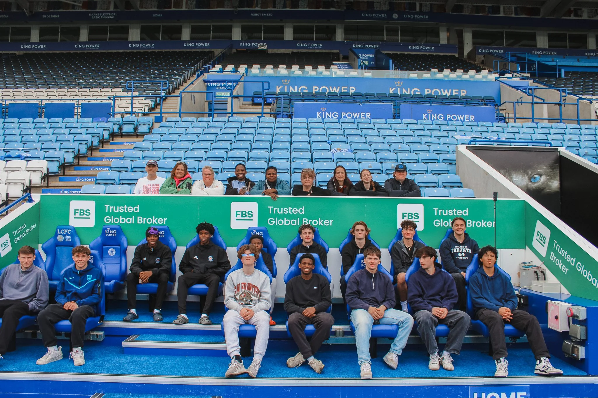 Group of young athletes and coaches sitting and standing on a sports bench inside a stadium, with many empty blue seats behind them and advertisements on the walls.
