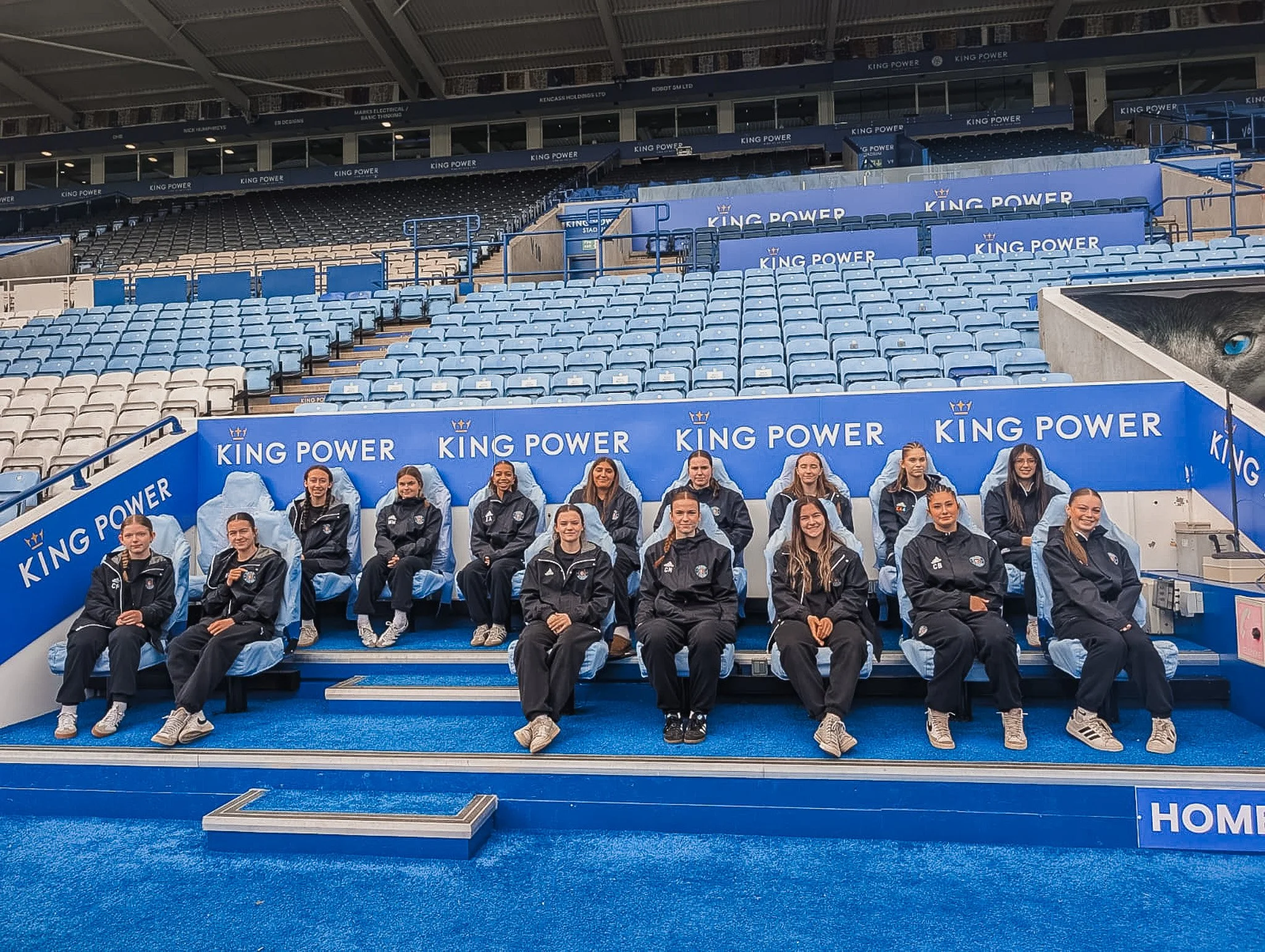 Group of fifteen young women in sportswear sitting on blue seats in a stadium, with 'KING POWER' banners in the background.