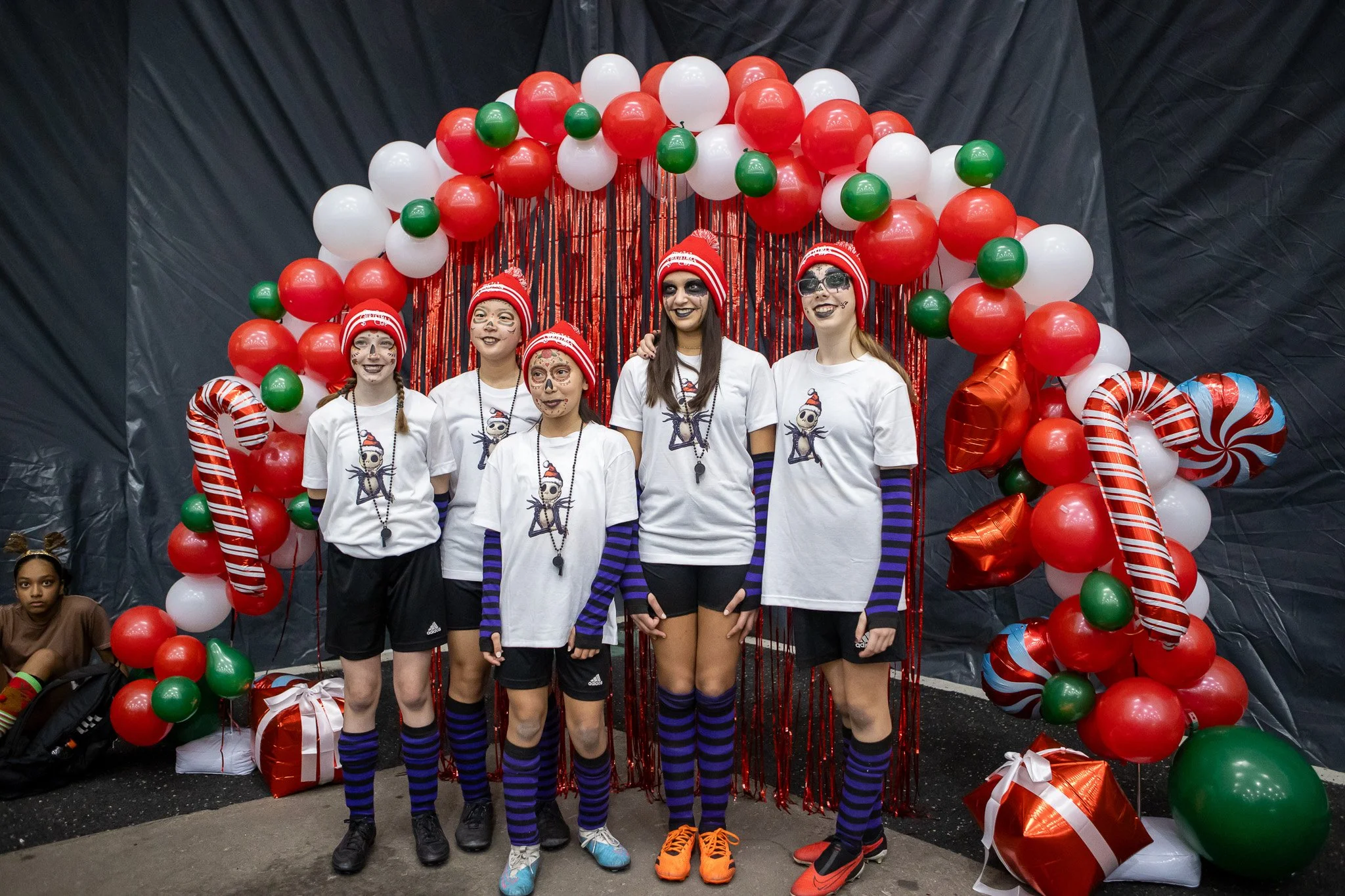 Group of five females dressed in skeleton-themed costumes with face paint, standing in front of a red, white, and green balloon arch with peppermint-shaped decorations and red foil wrapped gift boxes.