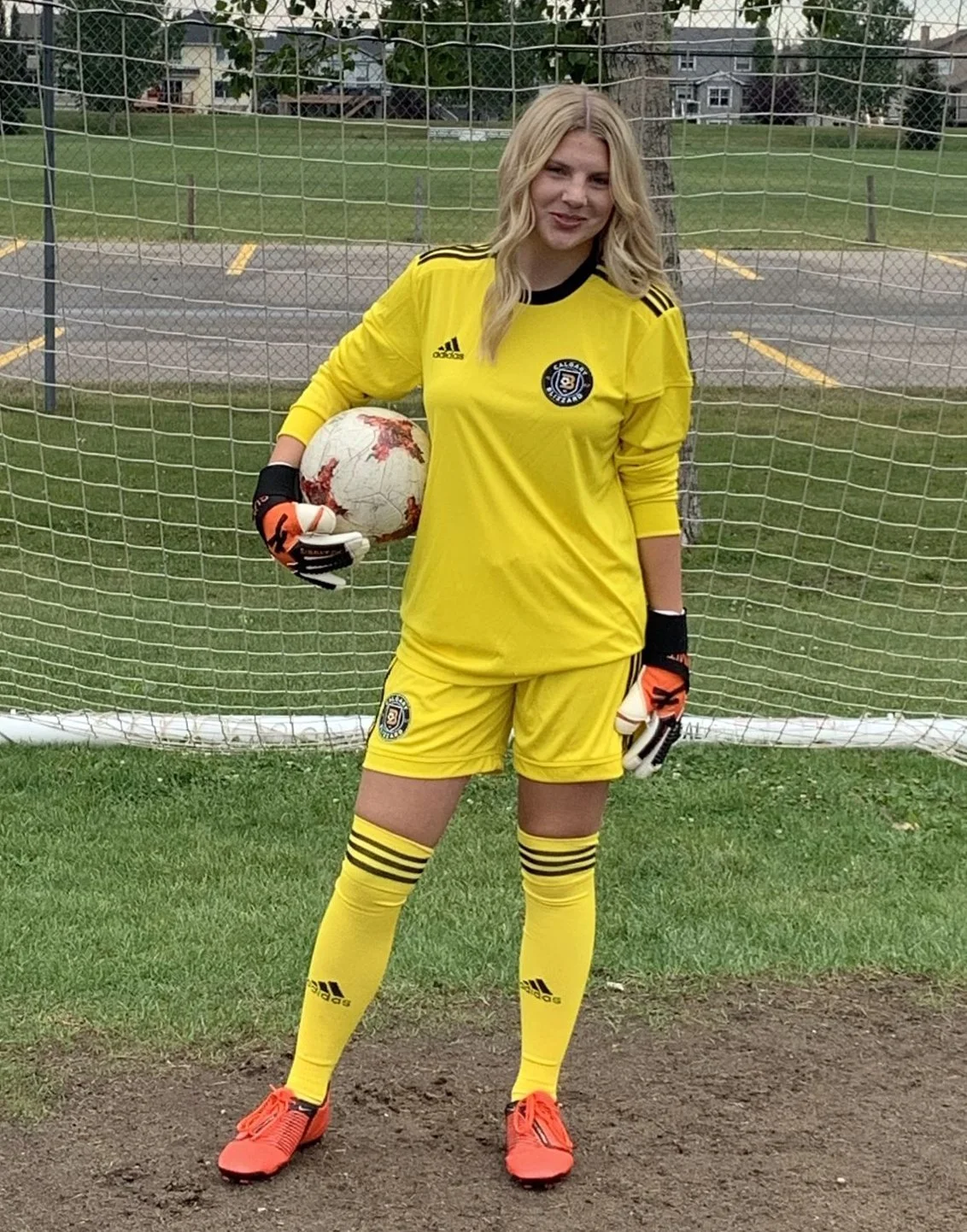 A female soccer player in yellow uniform standing in front of a goal, holding a soccer ball, with gloves and sports cleats, on grass near a parking lot.