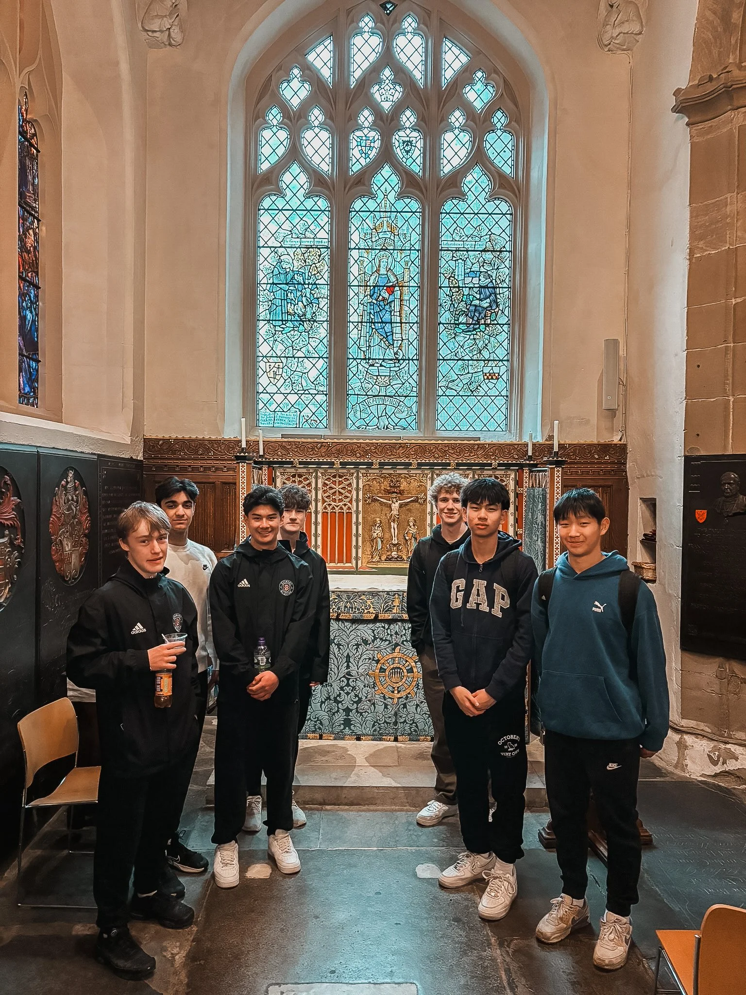 Group of seven boys standing inside a church near an altar with a colorful stained glass window behind them.