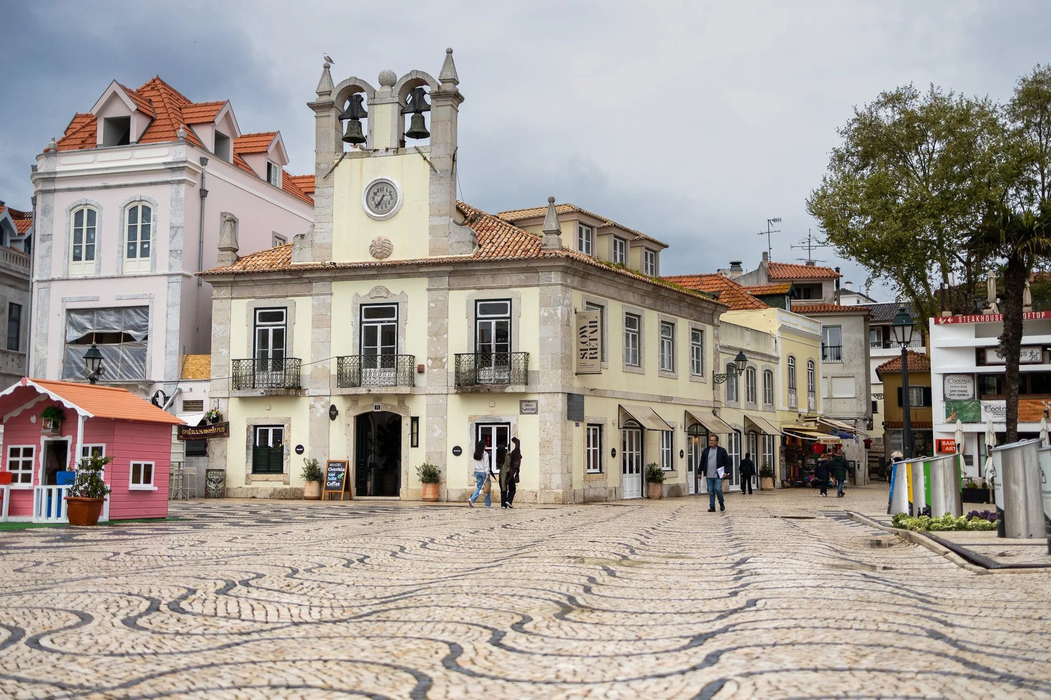 A historic building with a clock tower and small balconies, surrounded by shops and cafes, on a cobblestone street with a few pedestrians, under a cloudy sky.