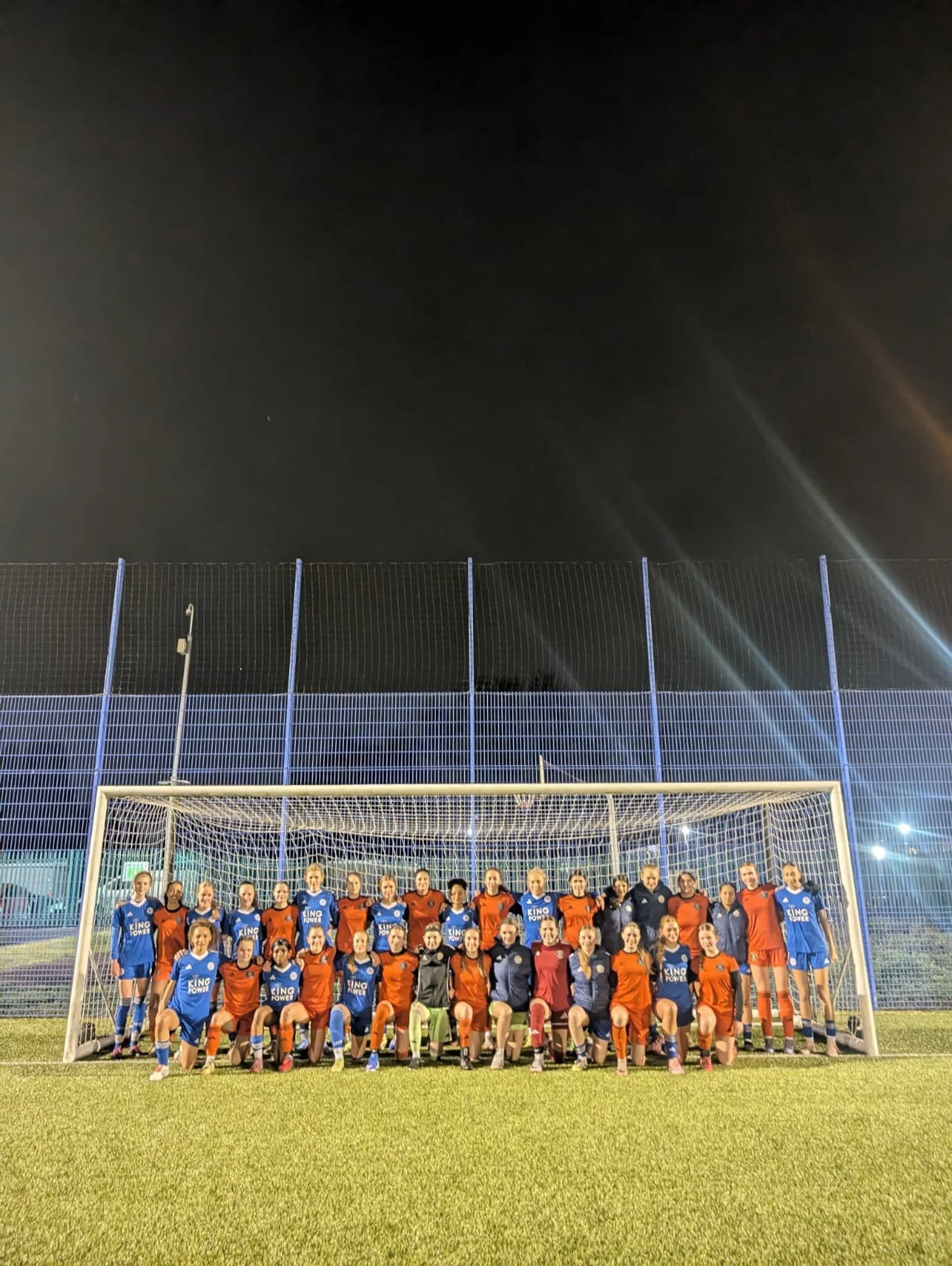 Group of female soccer players and coaches posing for a team photo in front of a goal on a soccer field at night, with a tall fence and dark sky in the background.