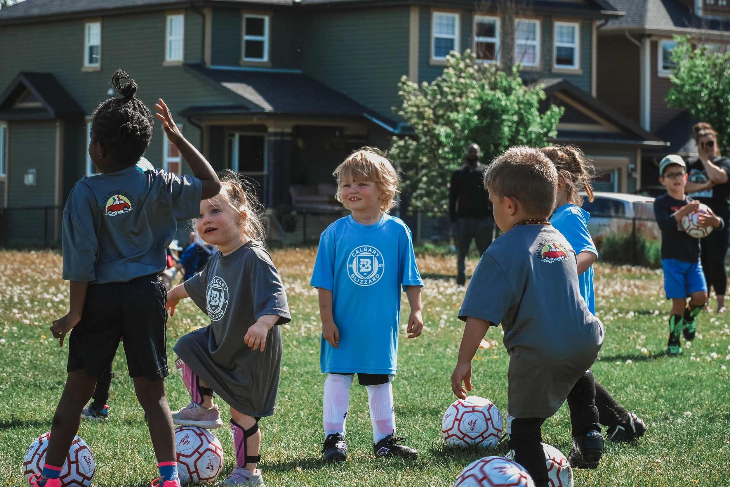 Children playing soccer on a grassy field on a sunny day, with some children holding soccer balls and others standing nearby.