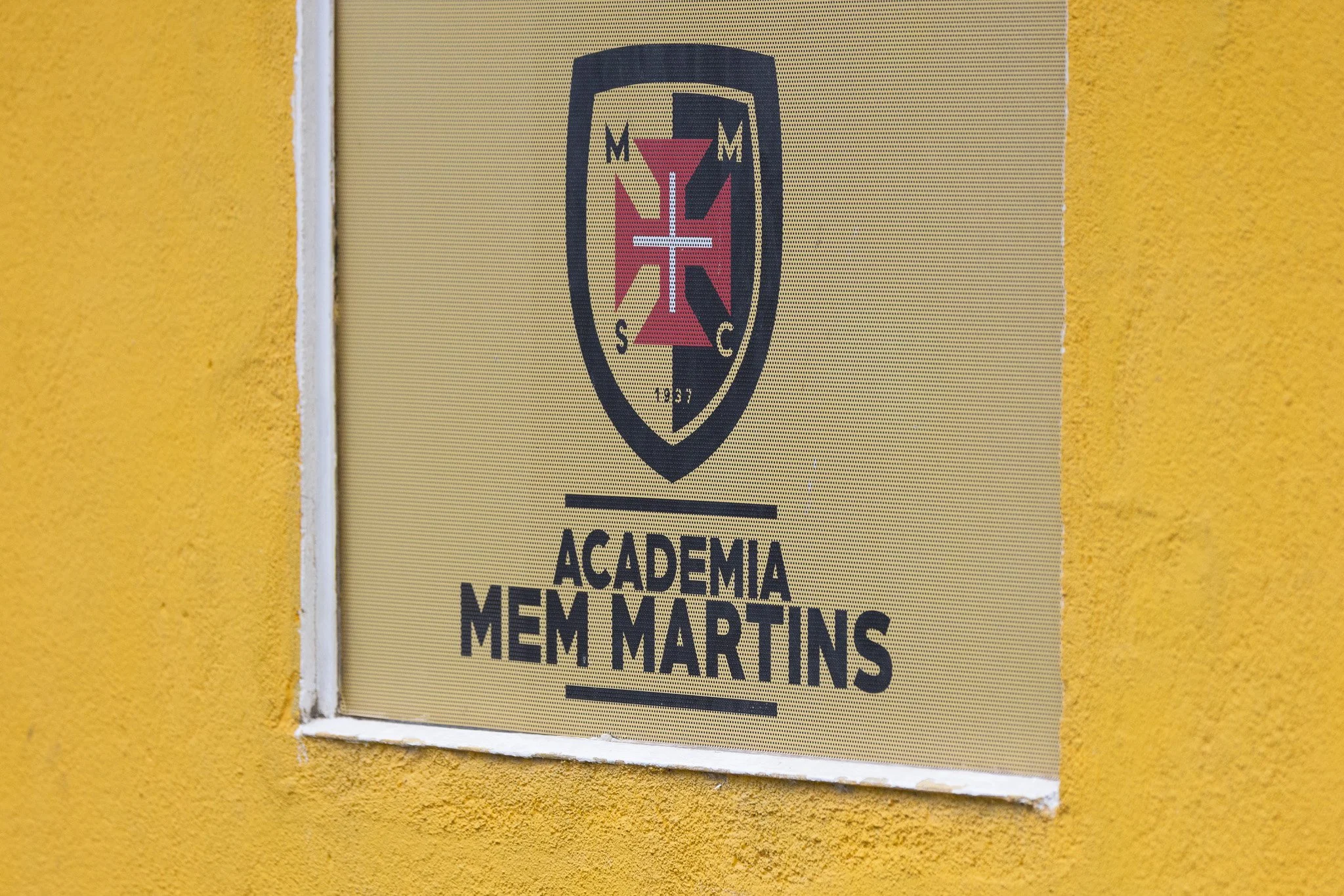 A sign on a yellow textured wall displaying the logo and name of Academia Mem Martins, featuring a shield emblem with a cross and the year 1957.