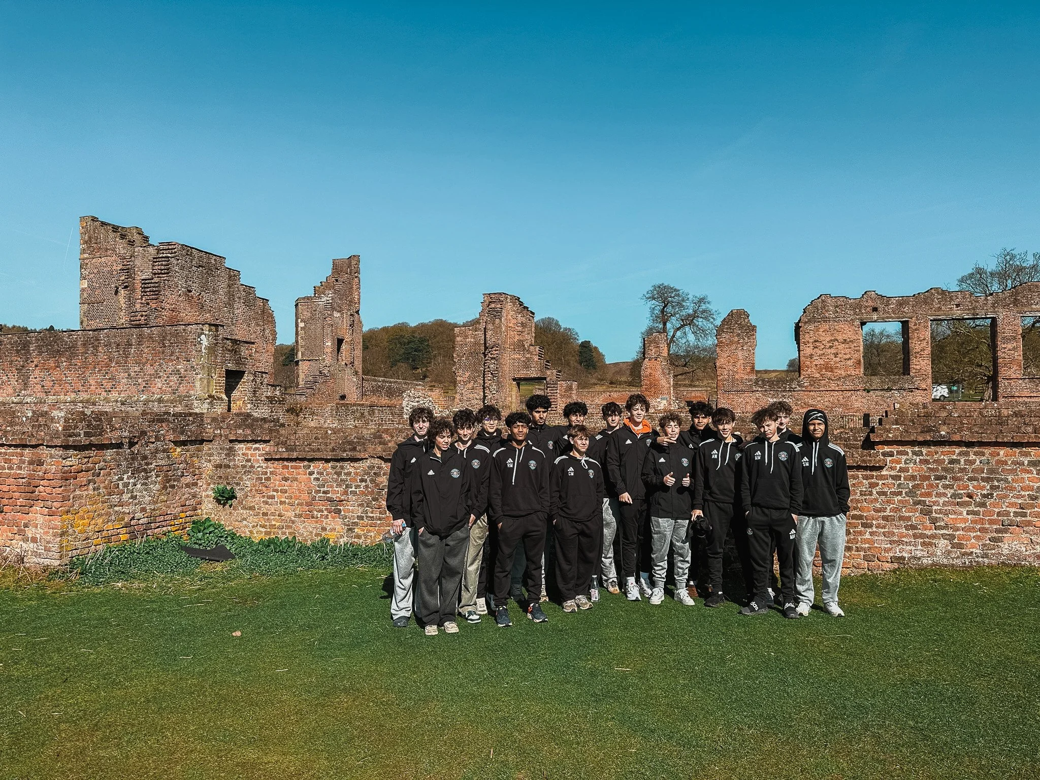Group of young soccer players in black sportswear standing on a grassy area with ruins of a brick building behind them, under a clear blue sky.